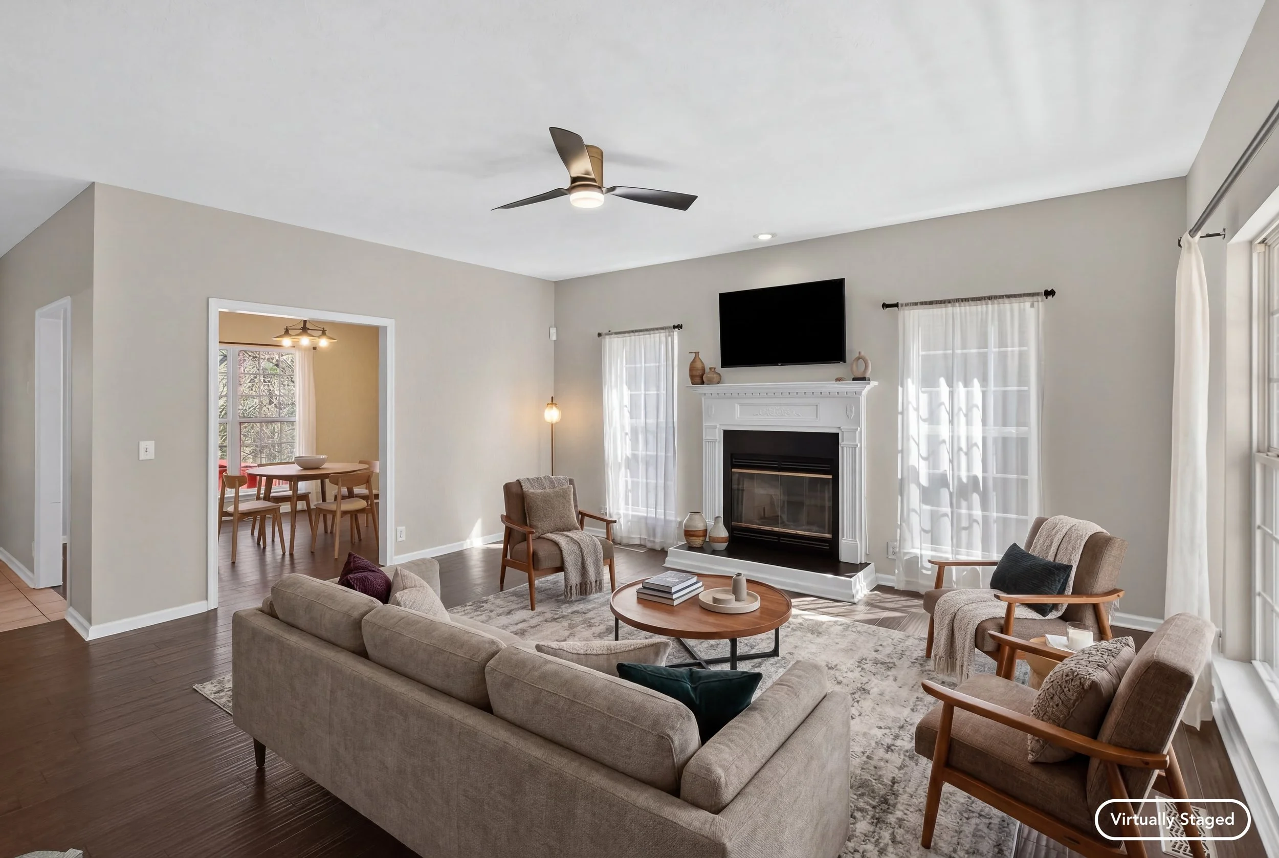 Living room with beige sofas, wooden chairs, a fireplace, and a TV mounted above the mantel. There are white curtains on the windows, and a rug covers part of the dark wooden floor. A small round coffee table has decorative items on it.