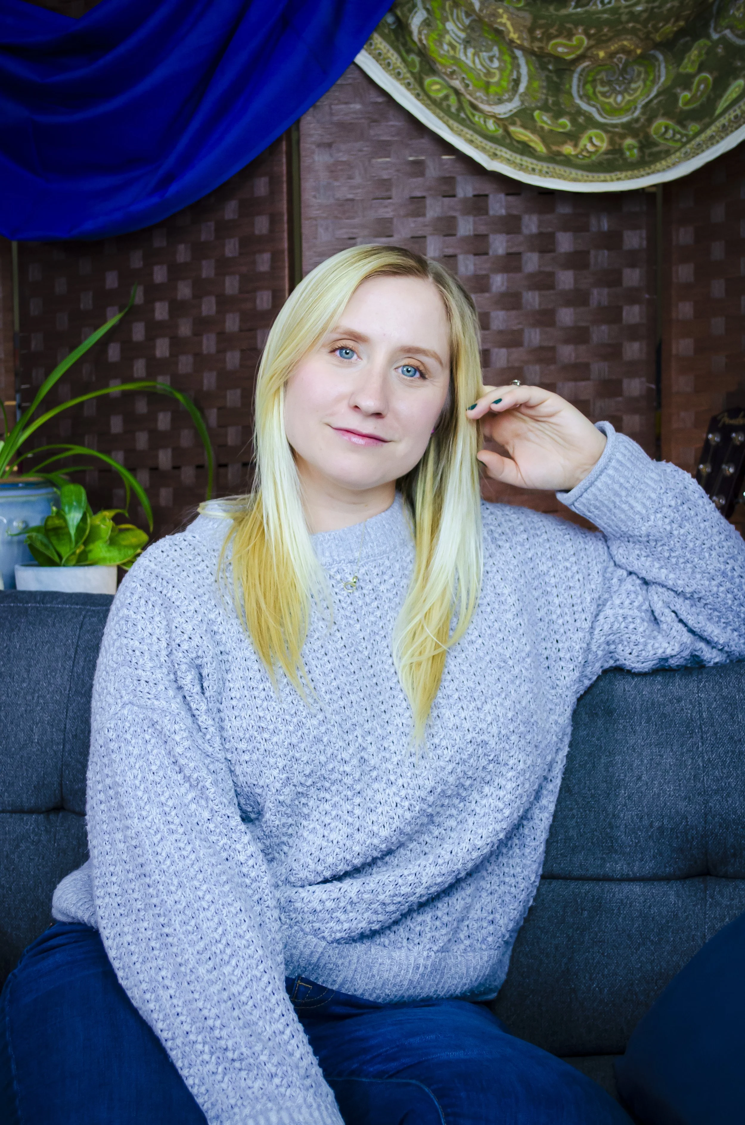 A young woman with long blonde hair, blue eyes, and fair skin, wearing a blue sweater, sitting on a dark gray couch in a room with a brown woven wall panel background, a blue curtain, and a decorative fabric piece hanging from the ceiling. She is smiling slightly and touching her hair with her right hand, with potted plants visible behind her.