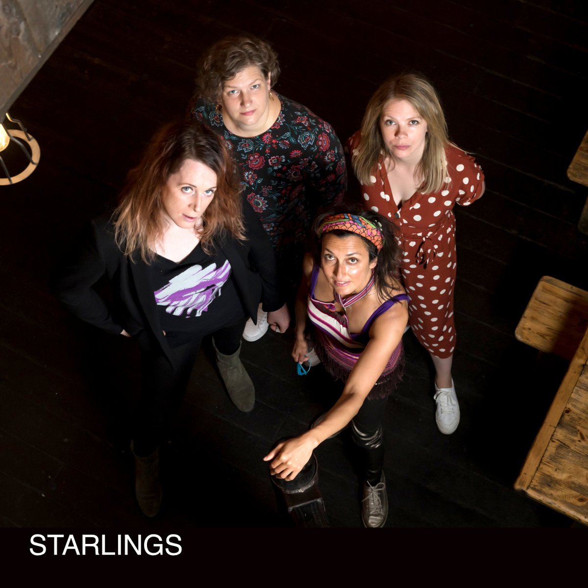 A group of four women (Starlings band from Bristol) looking up at the camera, standing on a dark wooden floor with wooden tables nearby.