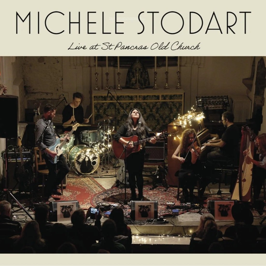 A music performance at St. Pancras Old Church featuring Michelle Stodart with a band, including drummer Emma Holbrook, guitarist David Ford, violinist Basia, Pianist, and a harpist, illuminated with warm string lights, audience in the foreground.