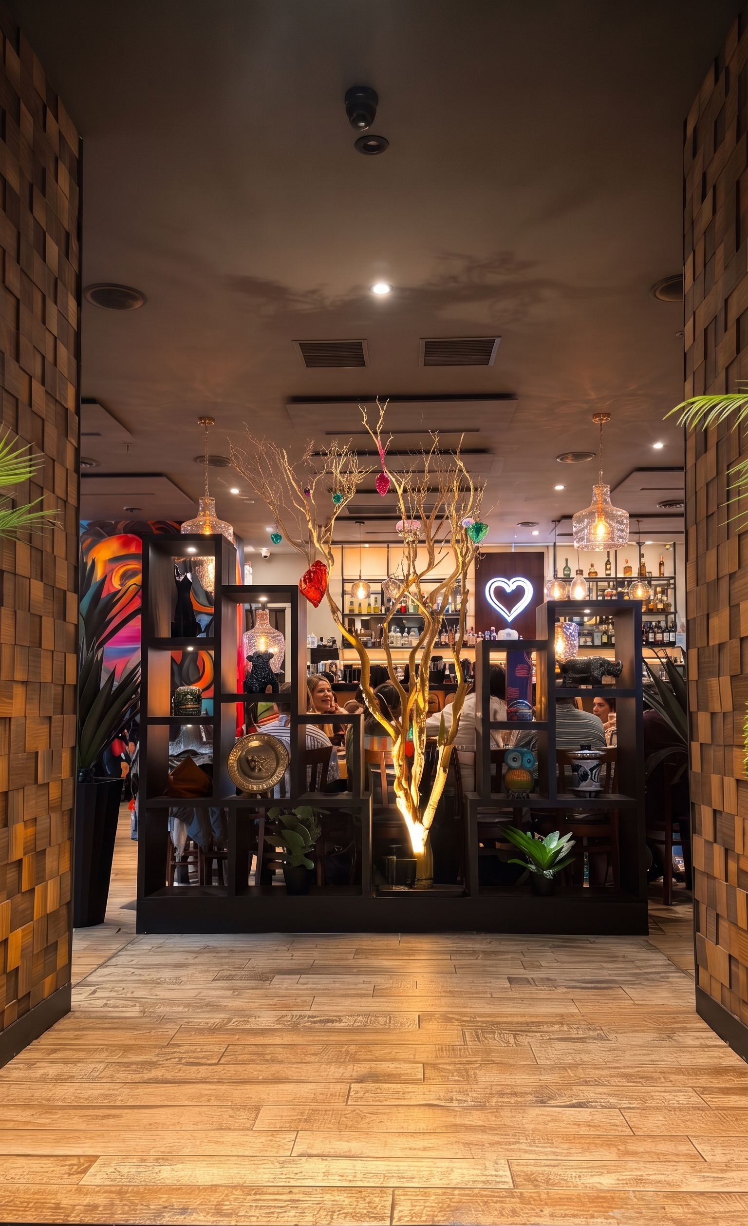 A cozy restaurant interior with wooden flooring, decorative partition with shelves and plants, illuminated bar area with a neon heart sign, and a central decorated tree with colorful ornaments.