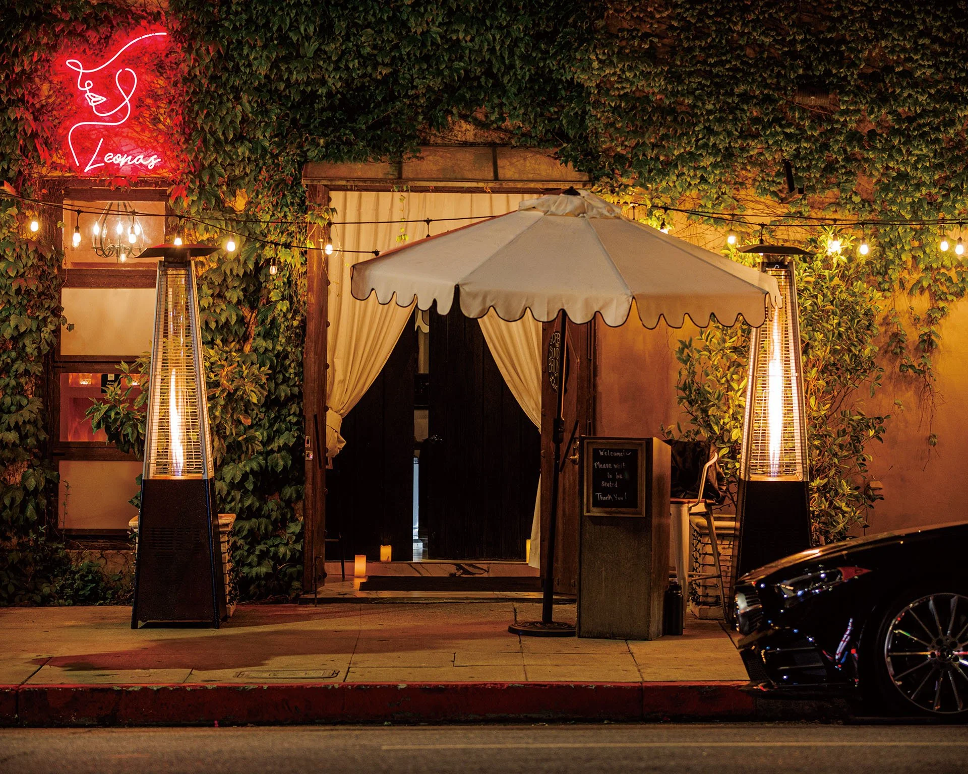 Nighttime exterior view of a restaurant or cafe entrance decorated with string lights and a large white patio umbrella. Green ivy covers the wall behind the entrance. Neon sign of a face with flowing hair and the name