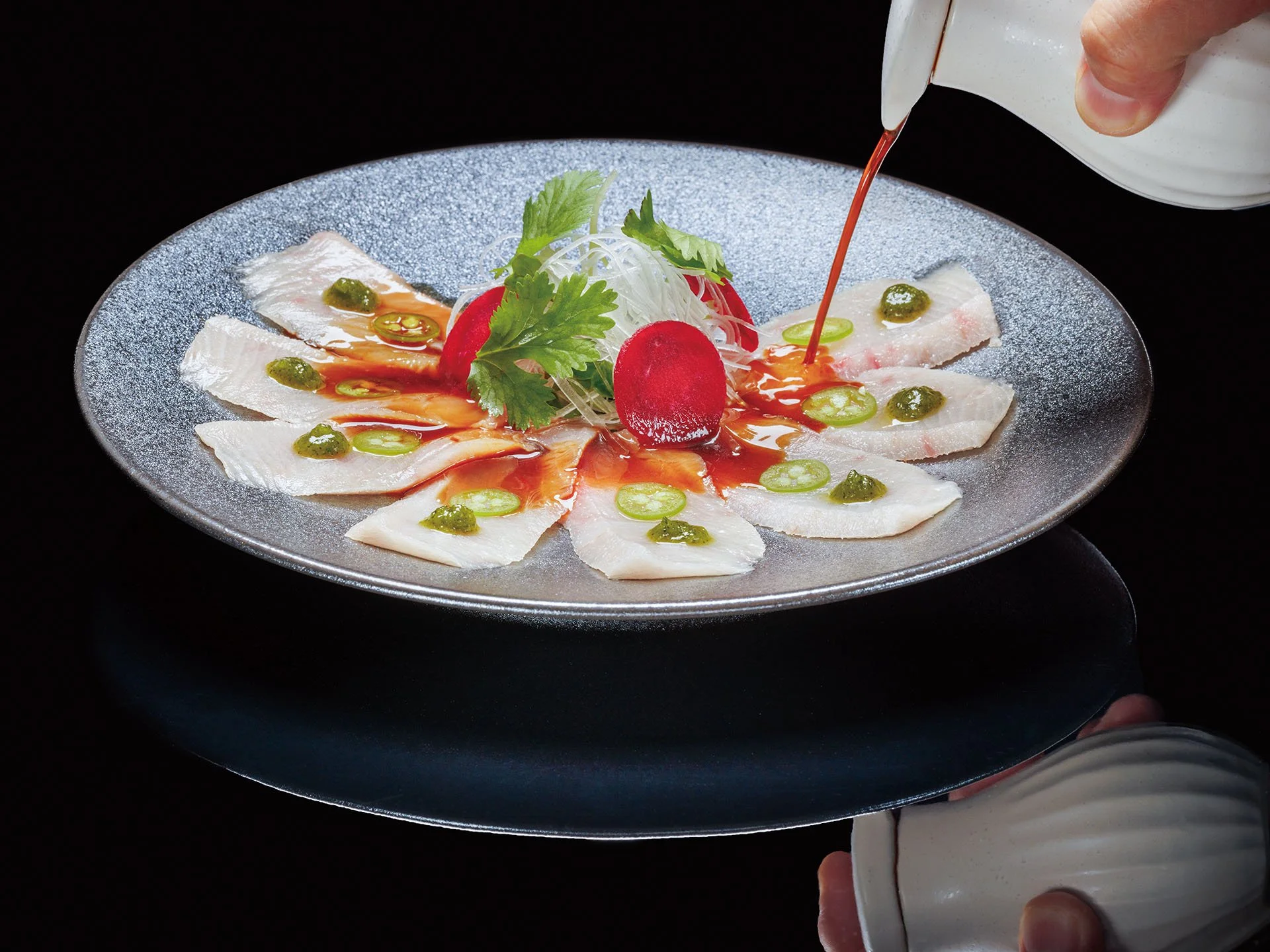 A chef pours sauce on a plate of sliced fish garnished with herbs, sliced chilies, and topped with radish and lettuce.