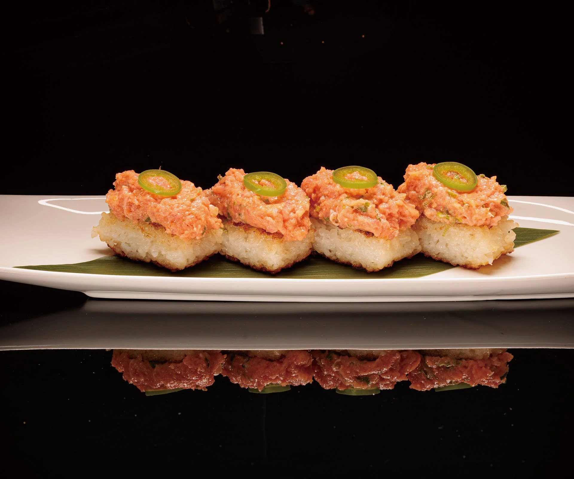 Close-up image of four pieces of sushi with a topping made of chopped raw fish and green chili slices, served on a white rectangular plate with a dark background.