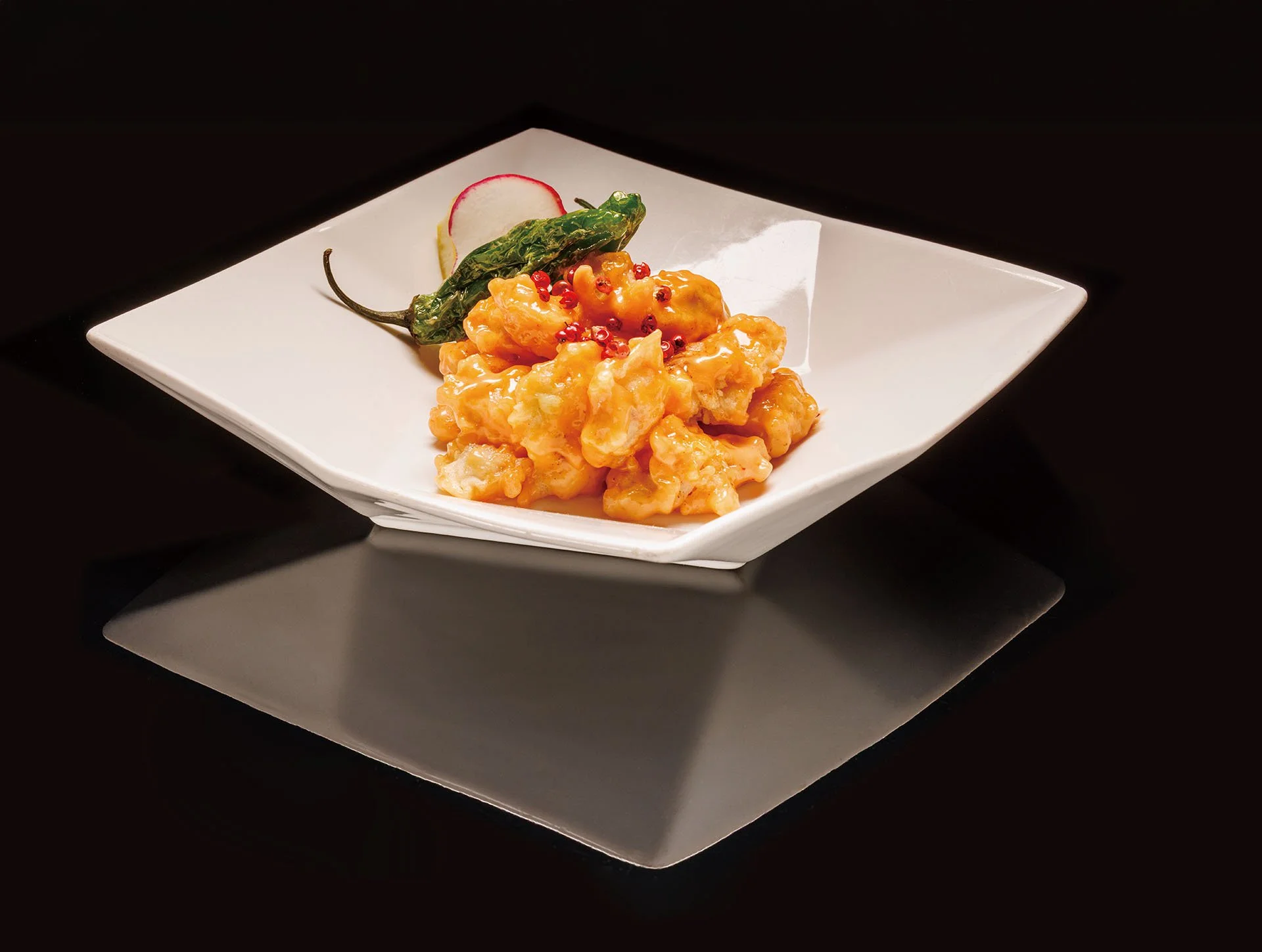 A plate of battered fried shrimp coated in orange sauce, garnished with pink peppercorns, a green chili pepper, a slice of radish, and a slice of radish, served on a white square dish with a black background.