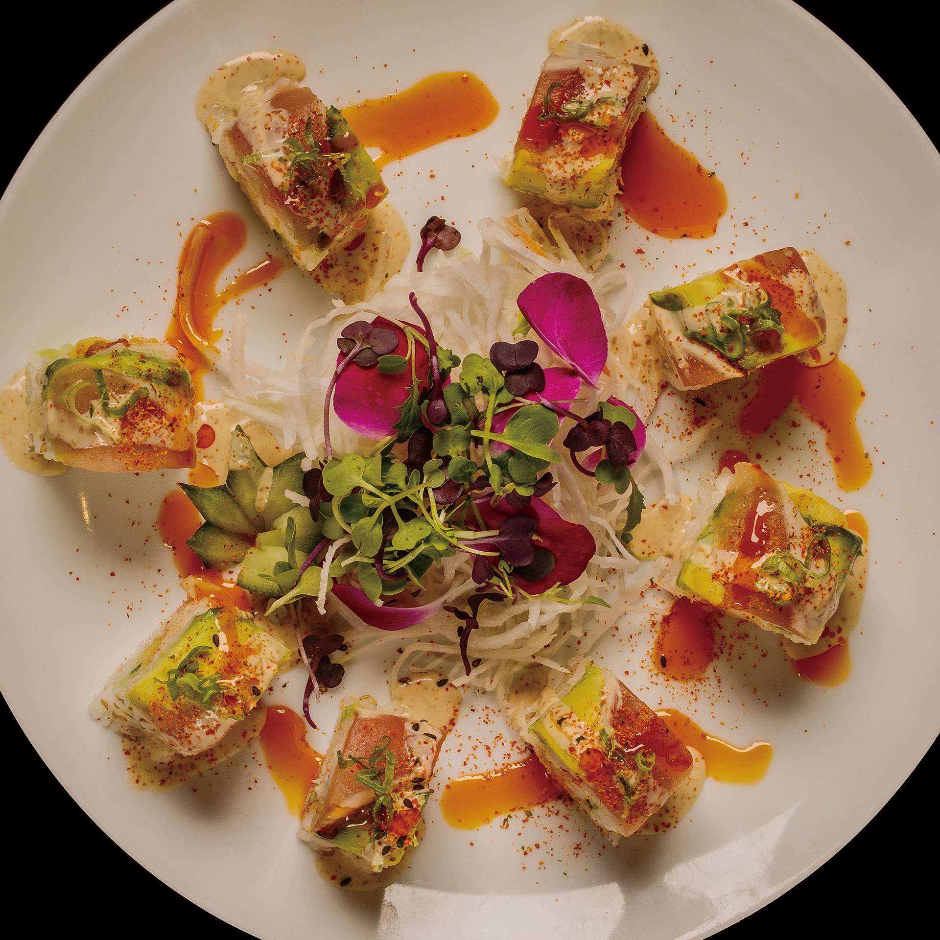 Sliced sushi rolls garnished with green onions and microgreens, drizzled with orange sauce, served with shredded vegetables and edible flowers on a white plate.