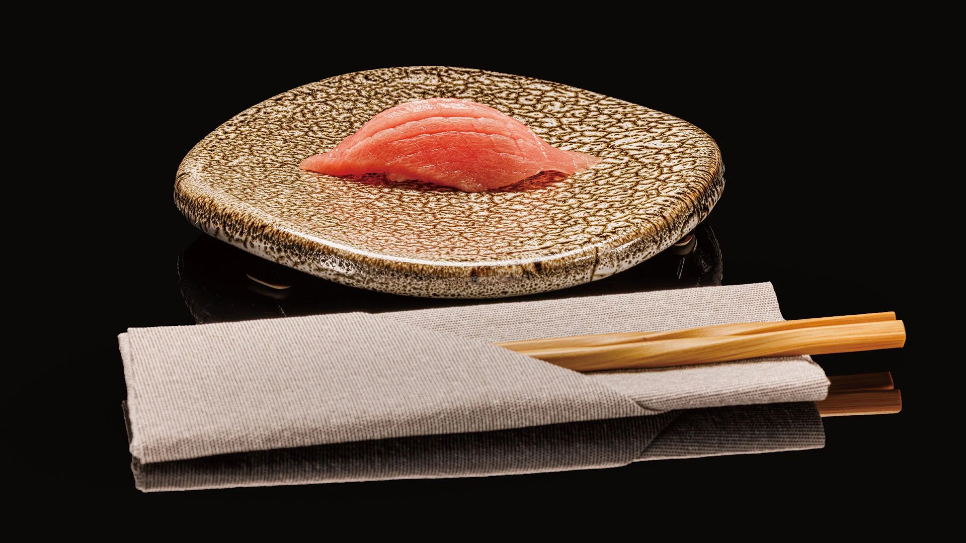 A single piece of Tuna sushi on a textured ceramic plate with chopsticks and a folded cloth napkin underneath. SUSHI BAR TETSUYA Sushi Row Book, preparing sushi in a restaurant or sushi bar. #SUSHIBARTEtsuya