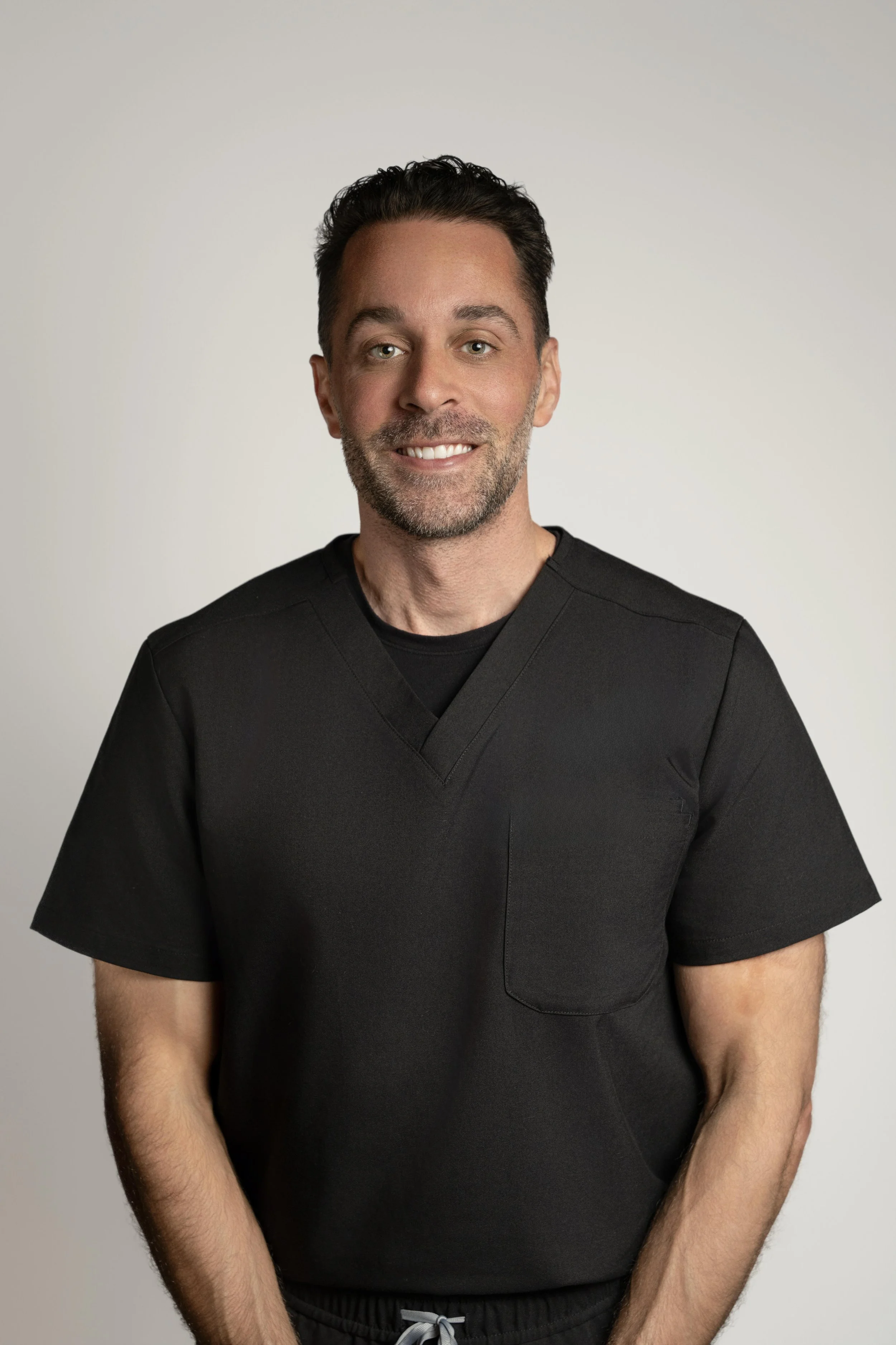 A man with dark hair, beard, and light skin, smiling in a black V-neck medical scrub top, standing against a plain light background.