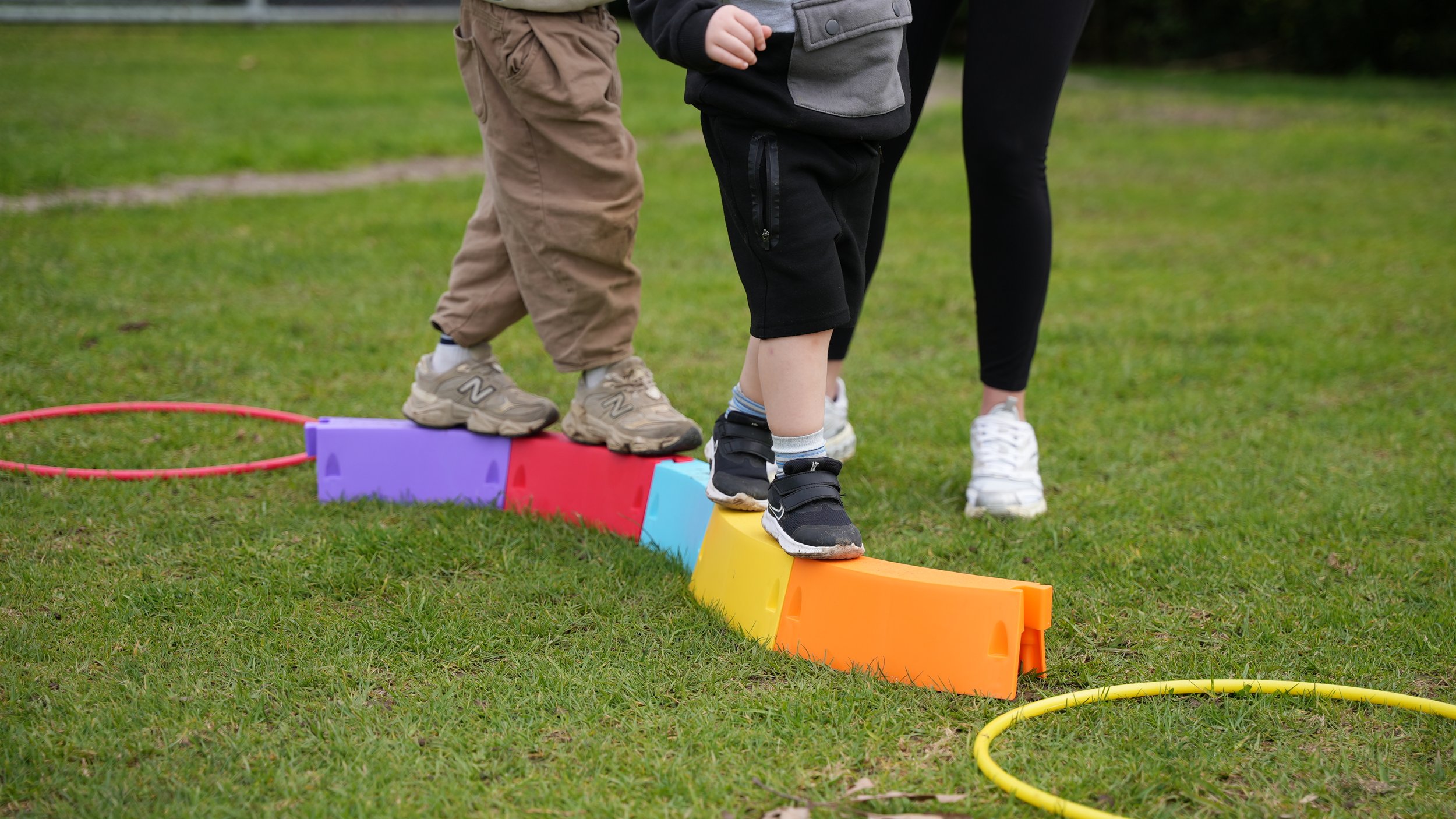 Children and an adult balancing on colorful stepping stones outdoors on a grassy field, with hula hoops and cones nearby, participating in an outdoor activity or game.