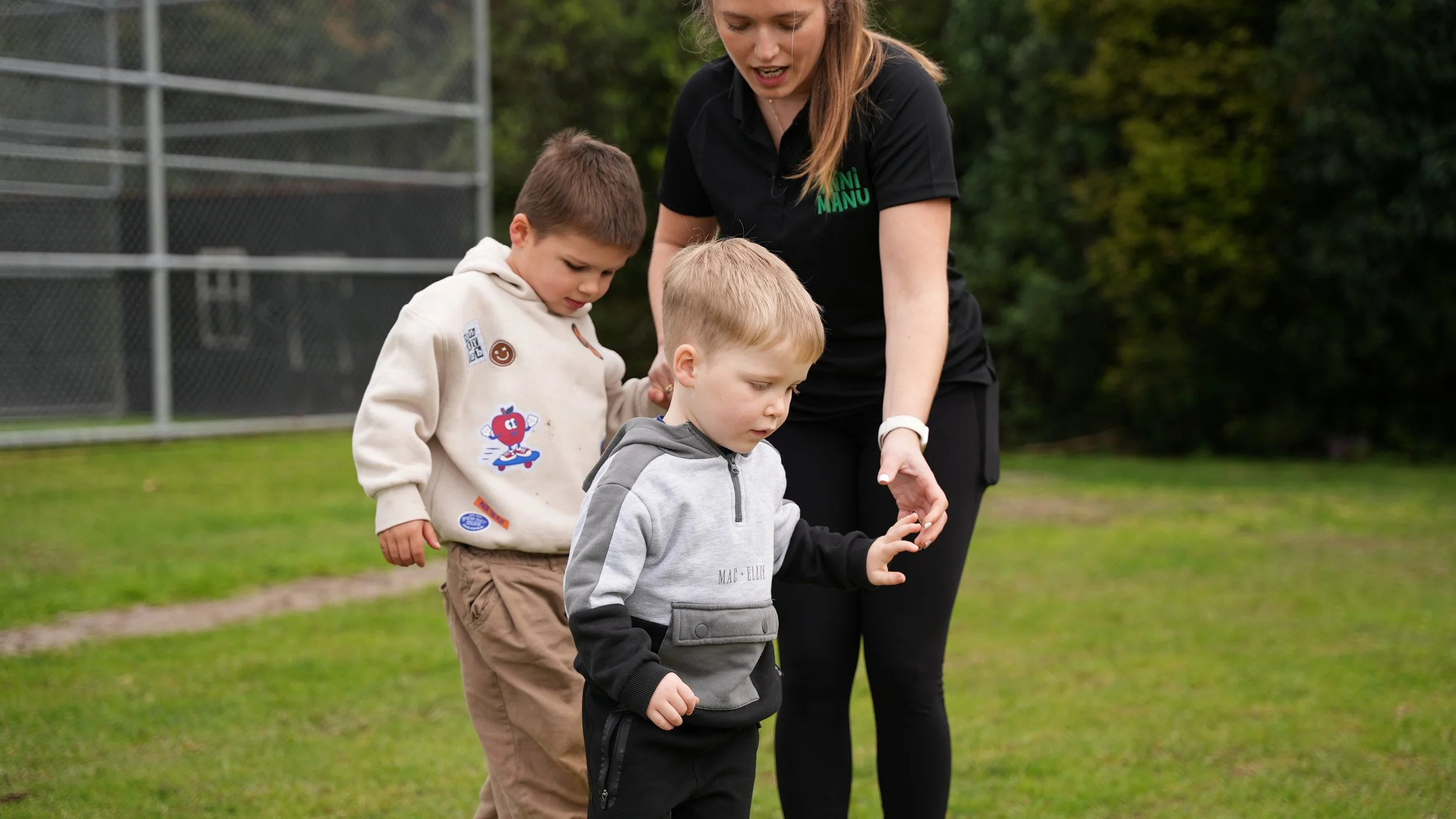 A woman with long hair in a black shirt guiding two young boys, one with light brown hair and one with darker hair, on a grassy field with a chain-link fence and trees in the background.
