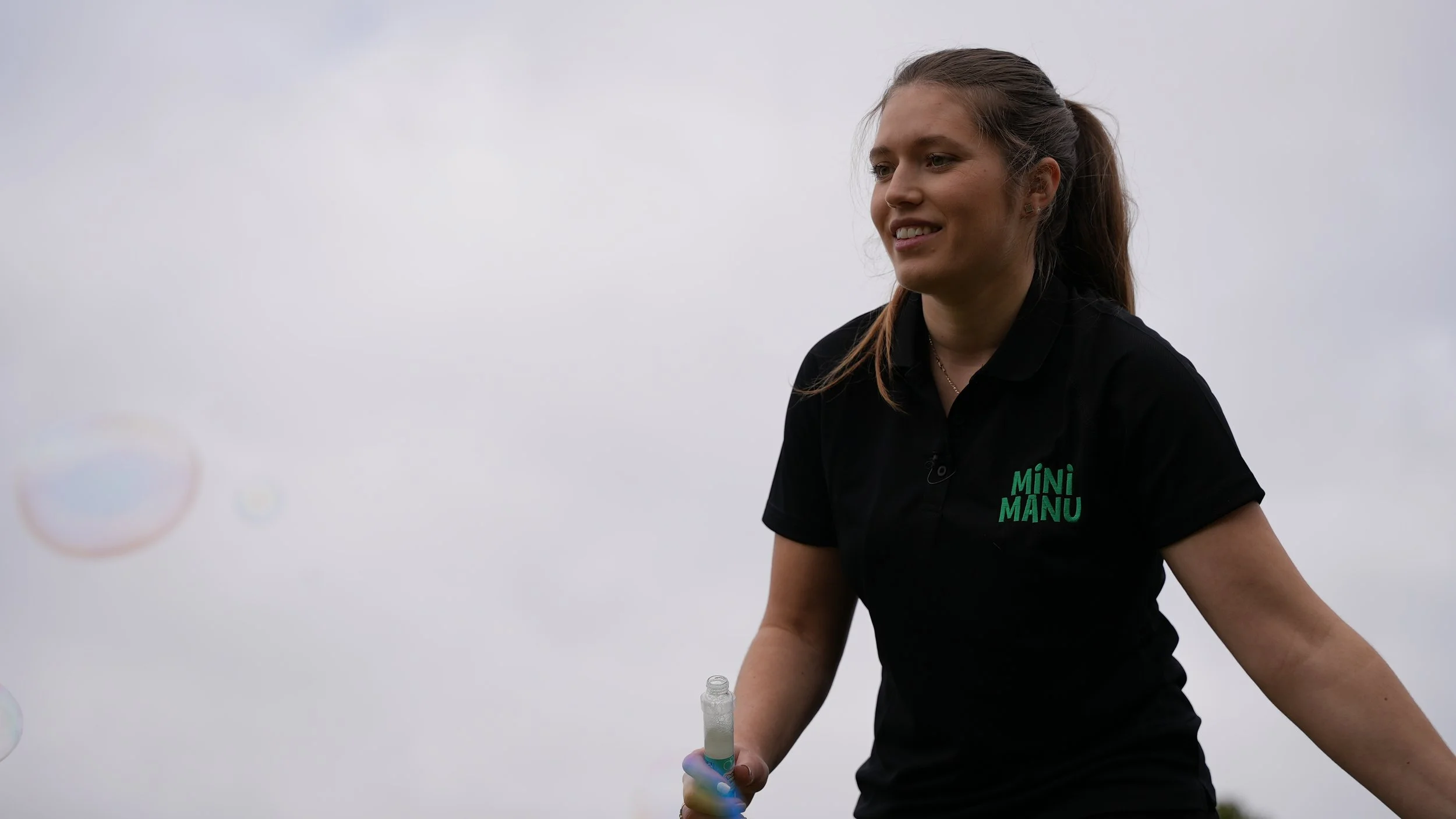 A young woman with a ponytail, wearing a black shirt with 'MINI MANU' printed on it, holding a bubble wand, outdoors against a cloudy sky.