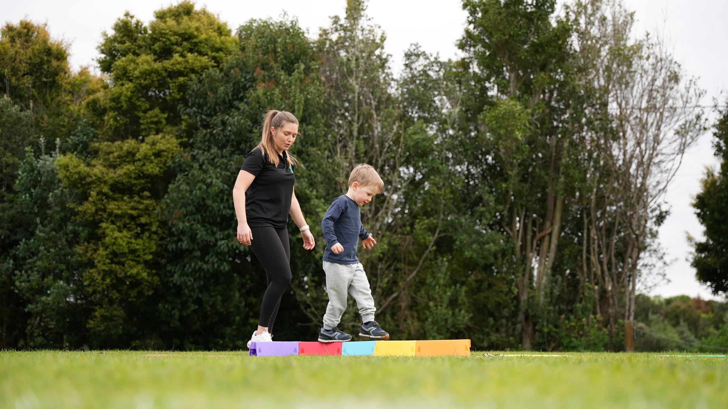 A woman helping a young boy walk across a colorful, rectangular stepping stone in a park with green trees in the background.