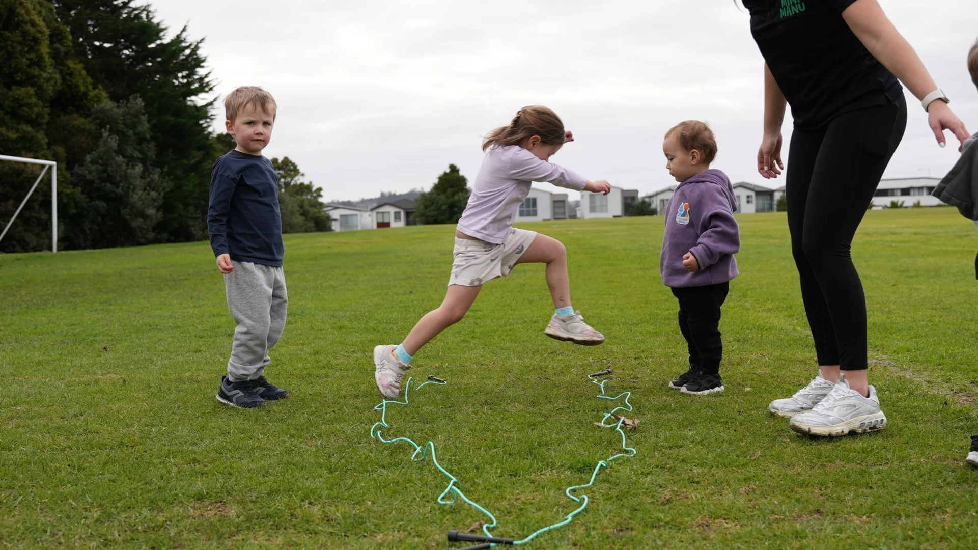 Children playing outdoor jump rope game, with two girls jumping and two boys standing nearby on a grassy field, with residential buildings and trees in the background.