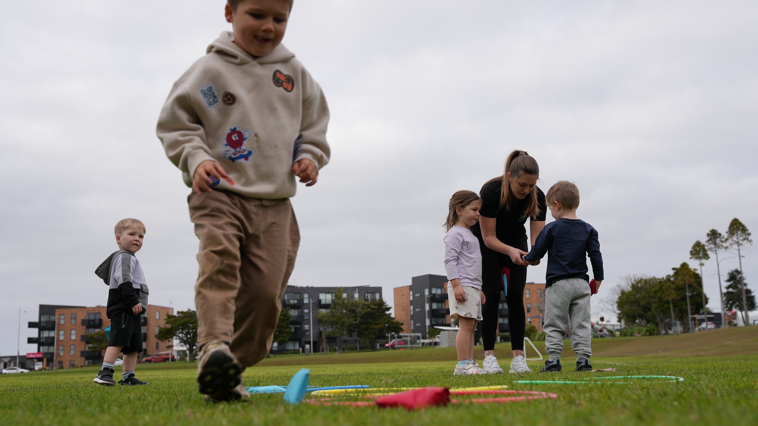 Children playing outdoor games on a grassy field with an adult supervising, in an urban area with apartment buildings in the background, under an overcast sky.