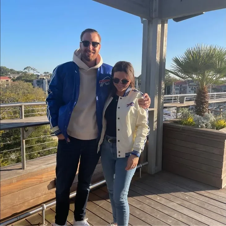 A man and woman wearing Buffalo Bills jackets, standing together outdoors on a balcony, with a view of trees and buildings in the background on a sunny day.