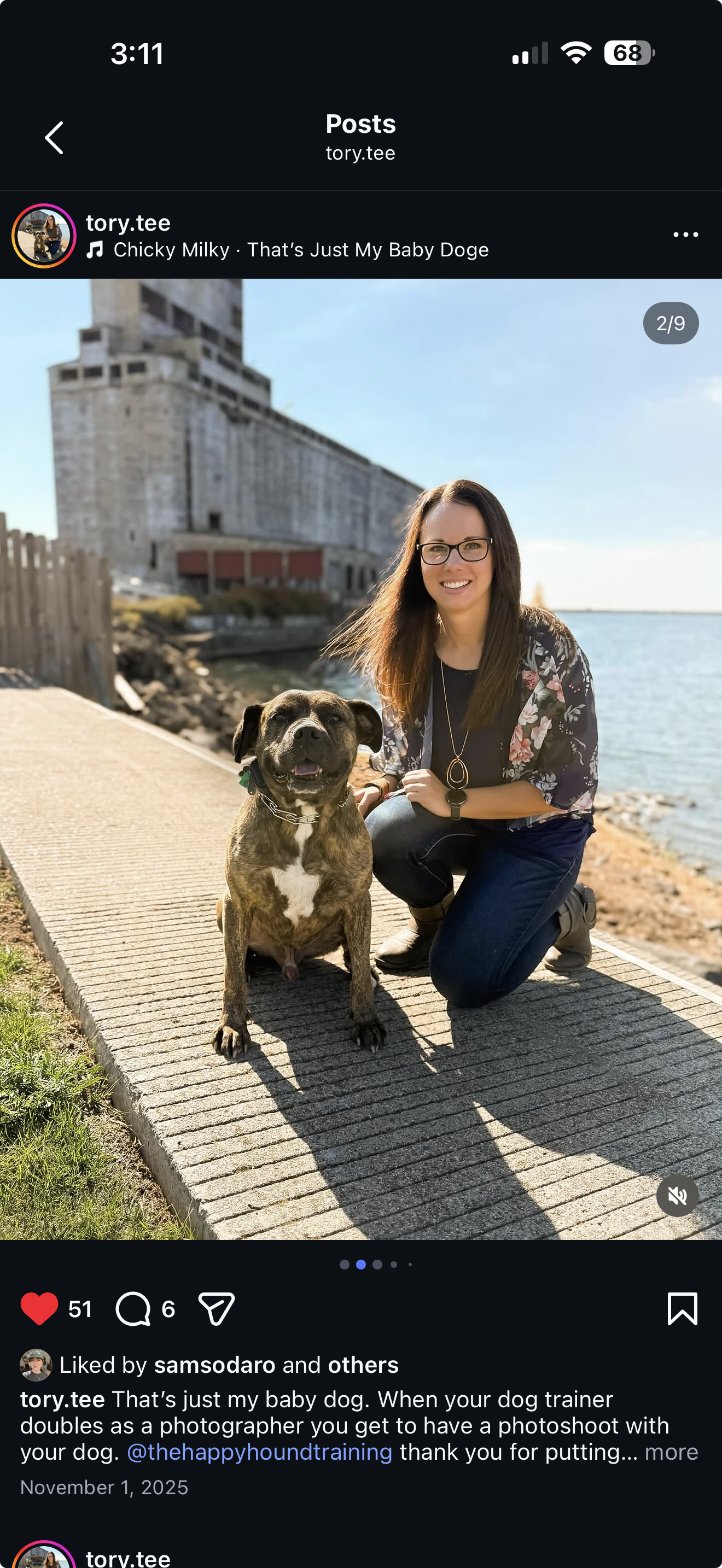 A woman with long brown hair, wearing glasses, a floral jacket, black top, and jeans, kneeling on a paved path beside a smiling brindle-colored dog with a white chest near a body of water and a large industrial building in the background.