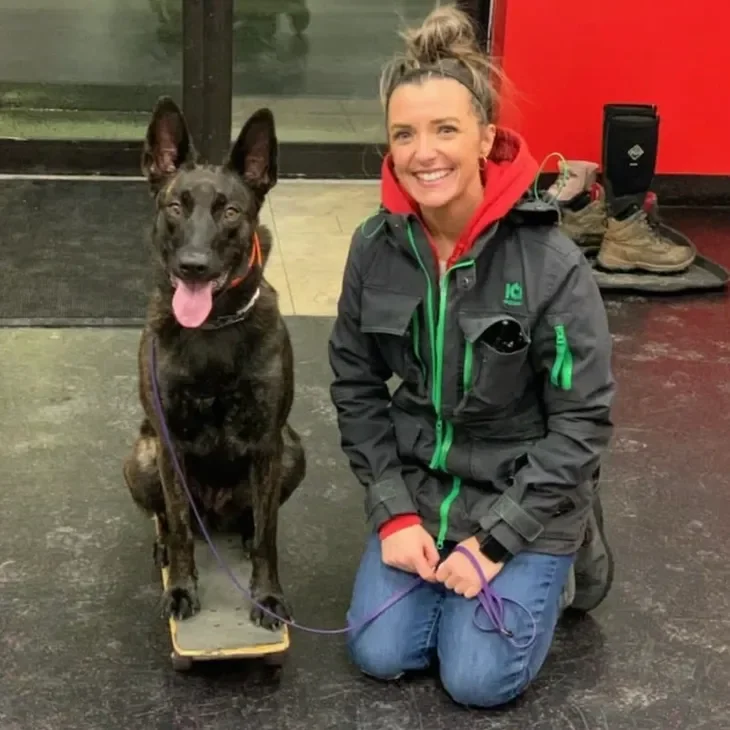 A smiling woman kneeling next to a large dog sitting on a skateboard indoors, with shoes and a bottle in the background.