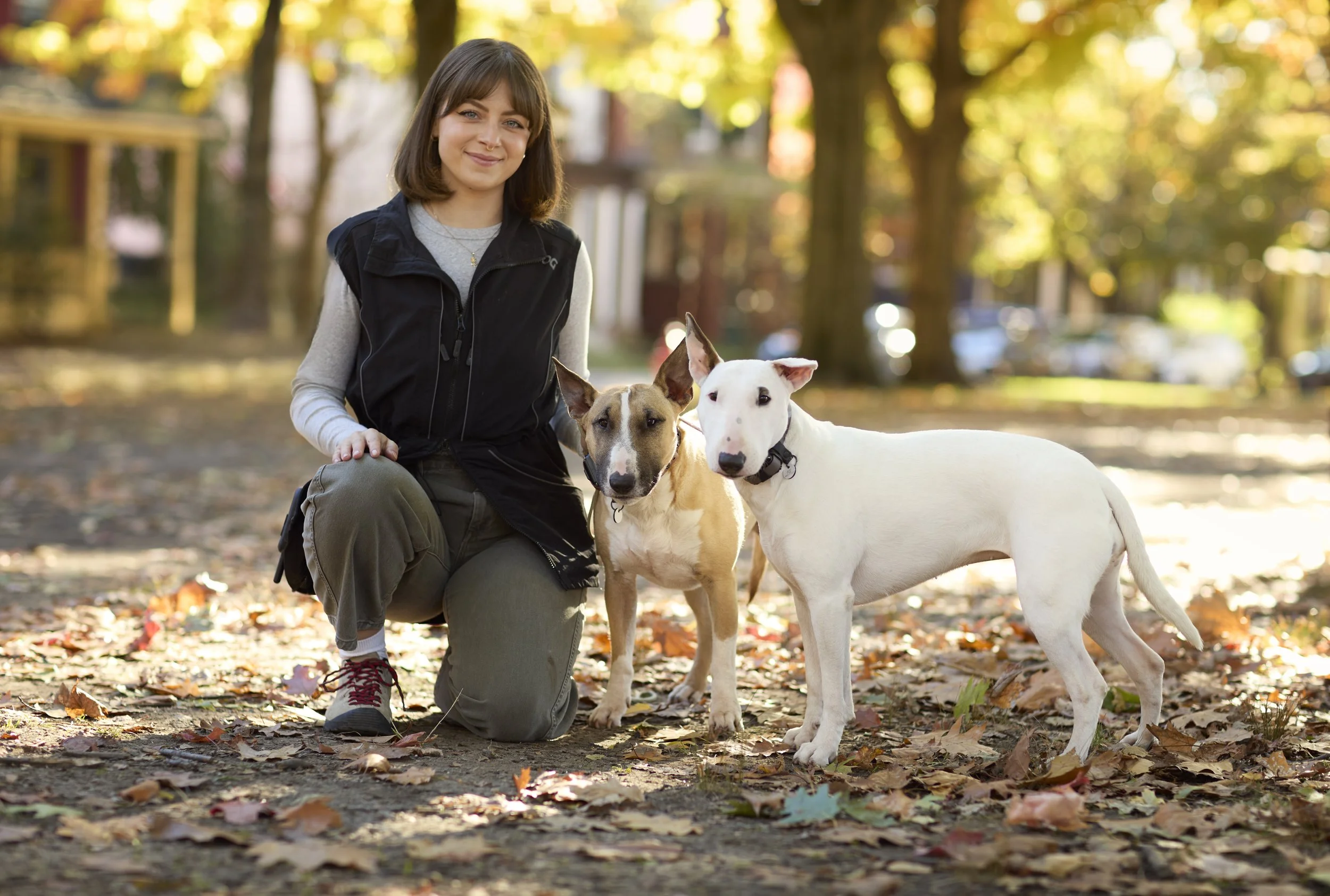 A woman with medium-length brown hair kneeling outdoors with two dogs on a leaf-covered ground during fall, in a park with trees and a blurred background.