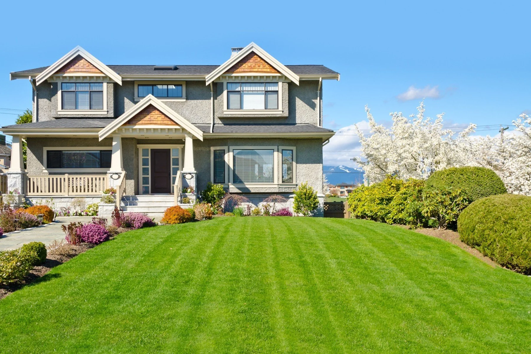 A large, two-story house with a well-manicured lawn, colorful flower beds, and bushes, set against a blue sky with white clouds and blooming trees.