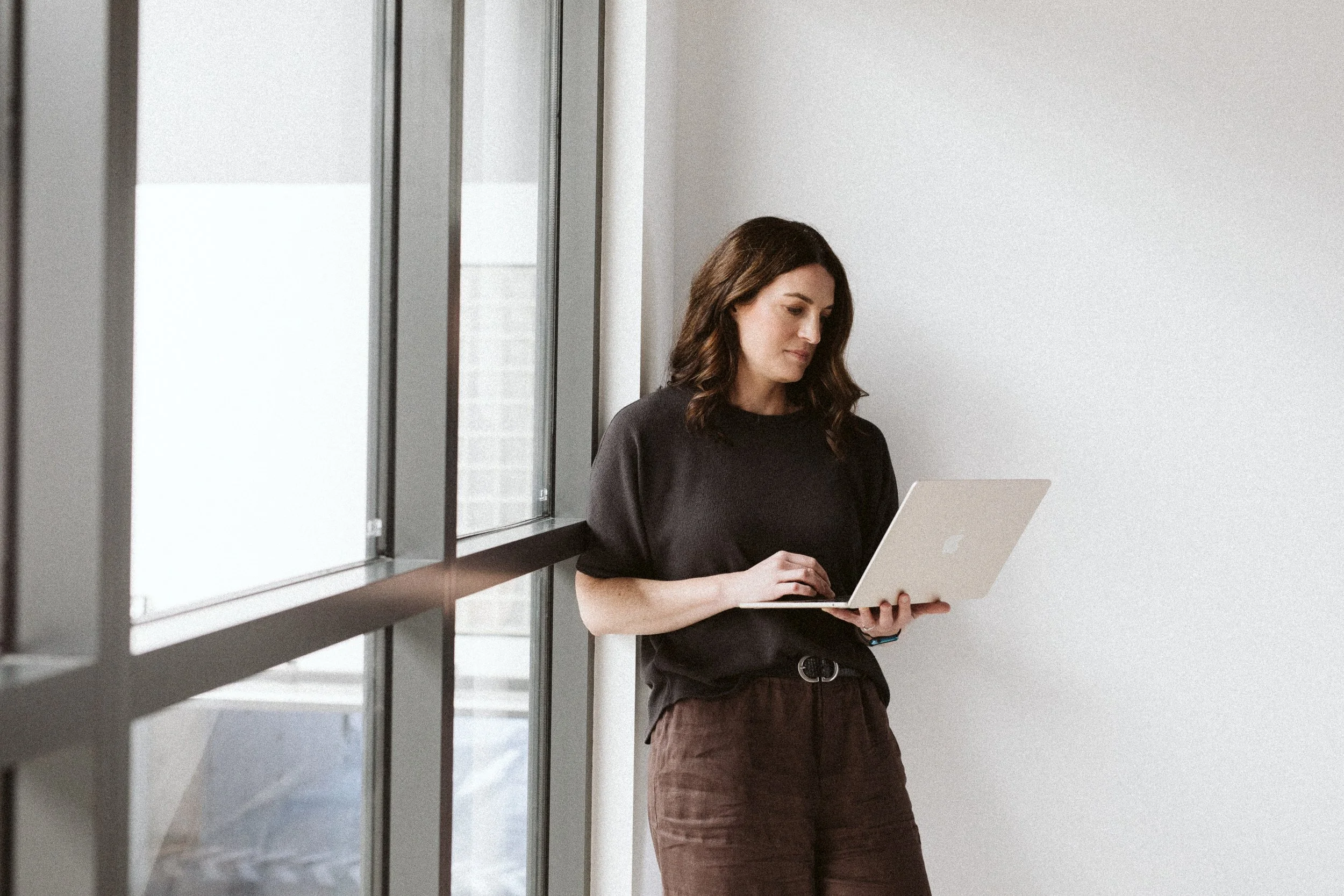 A woman with brown, wavy hair is standing by a large window on a white wall, working on a silver laptop, dressed in a black top and brown pants in a modern office or home setting.