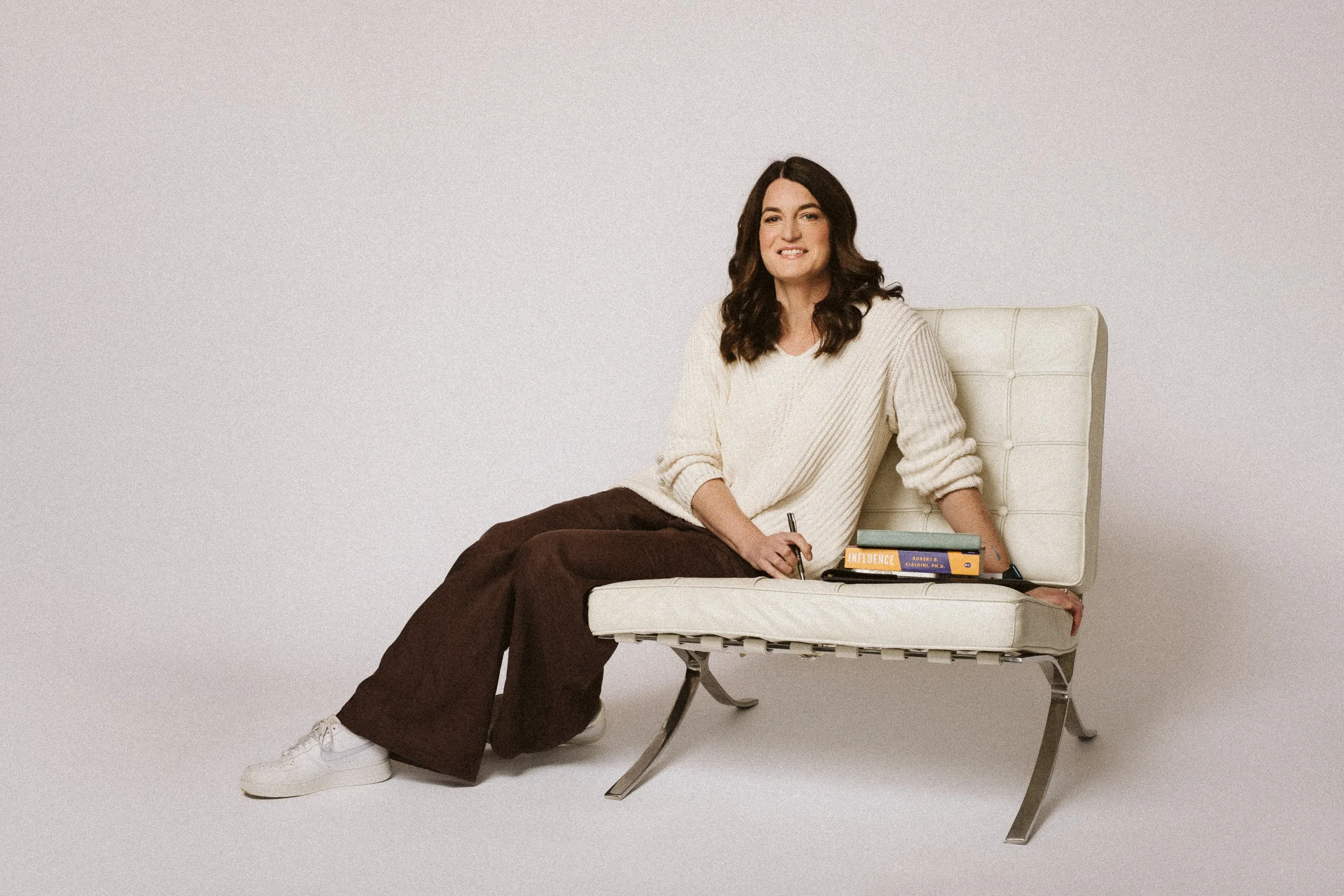 A woman sitting on a white lounge chair, holding a pen and a stack of books, with a plain light background.