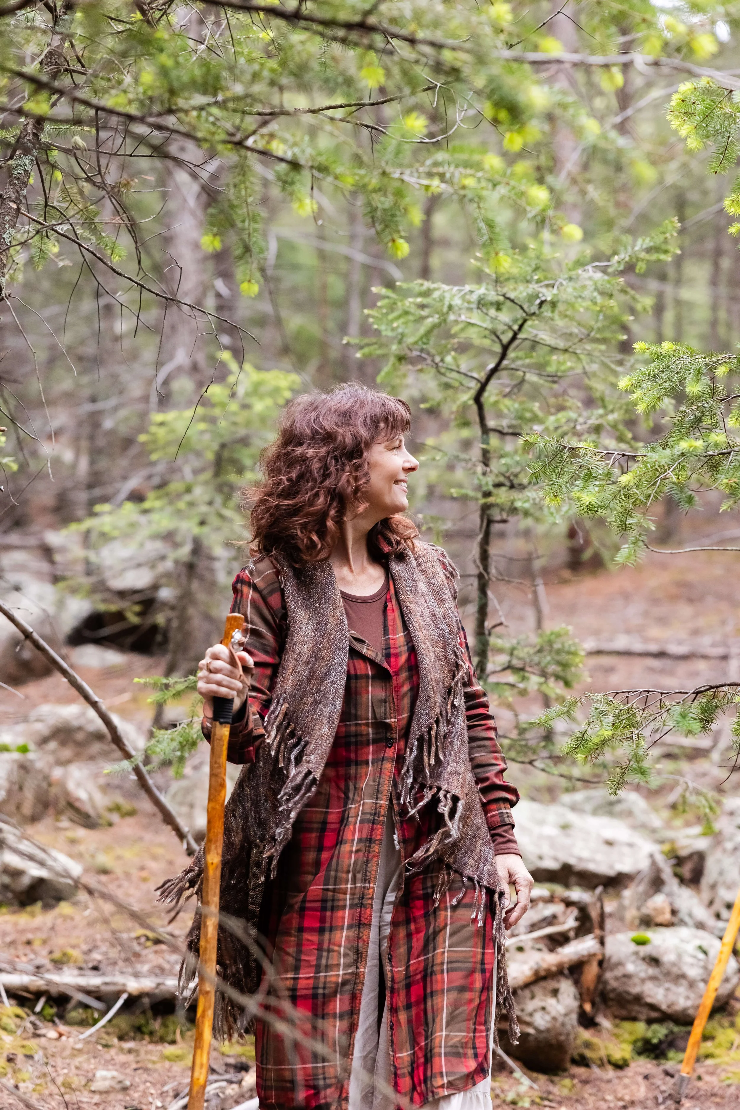 A woman with curly brown hair in Colorado, smiling, wearing a plaid shirt and a fringed vest, holding a walking stick.