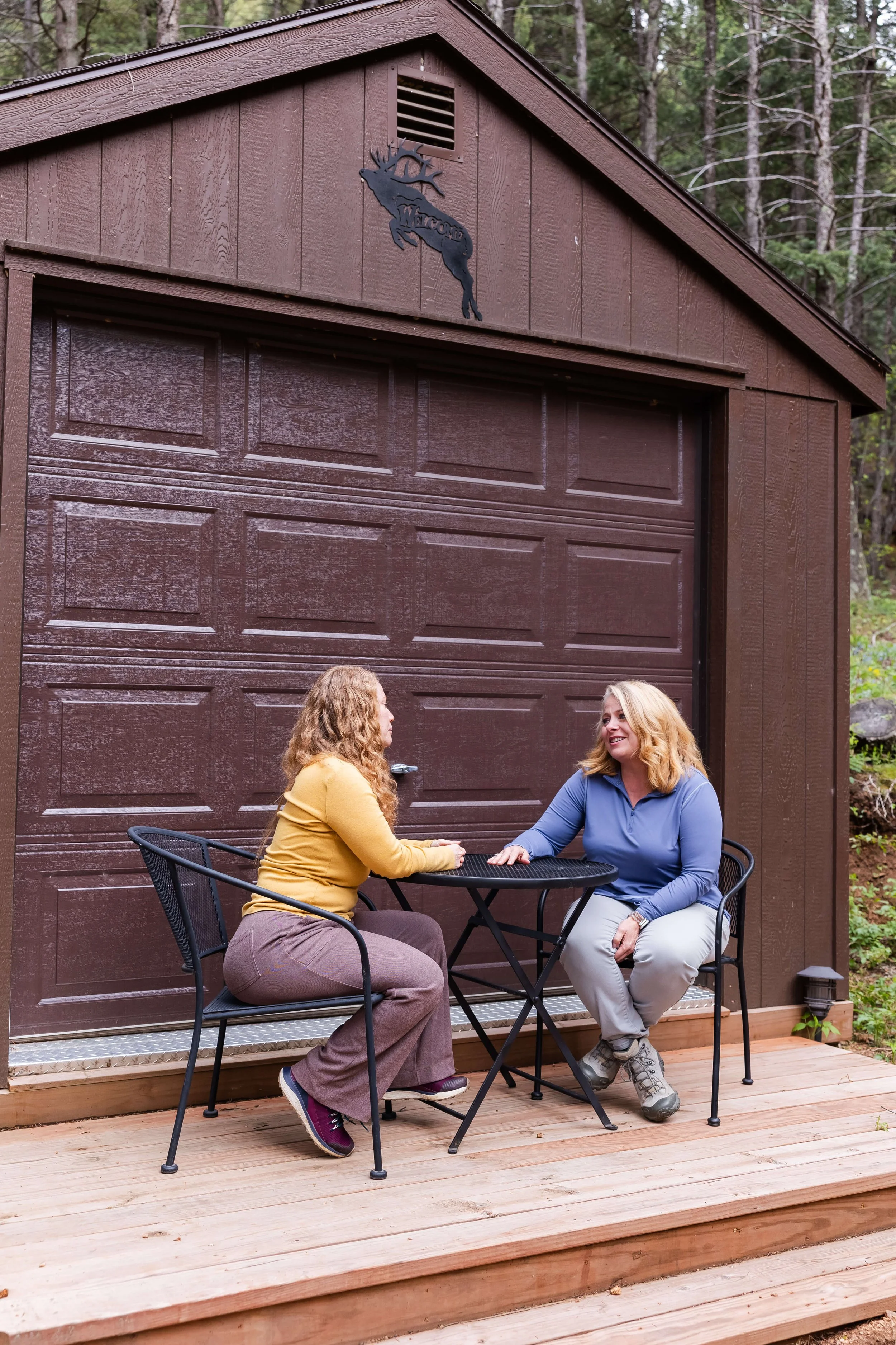 Two women are sitting at a black patio table on a wooden deck in front of a brown garage at a mountain wellness retreat.