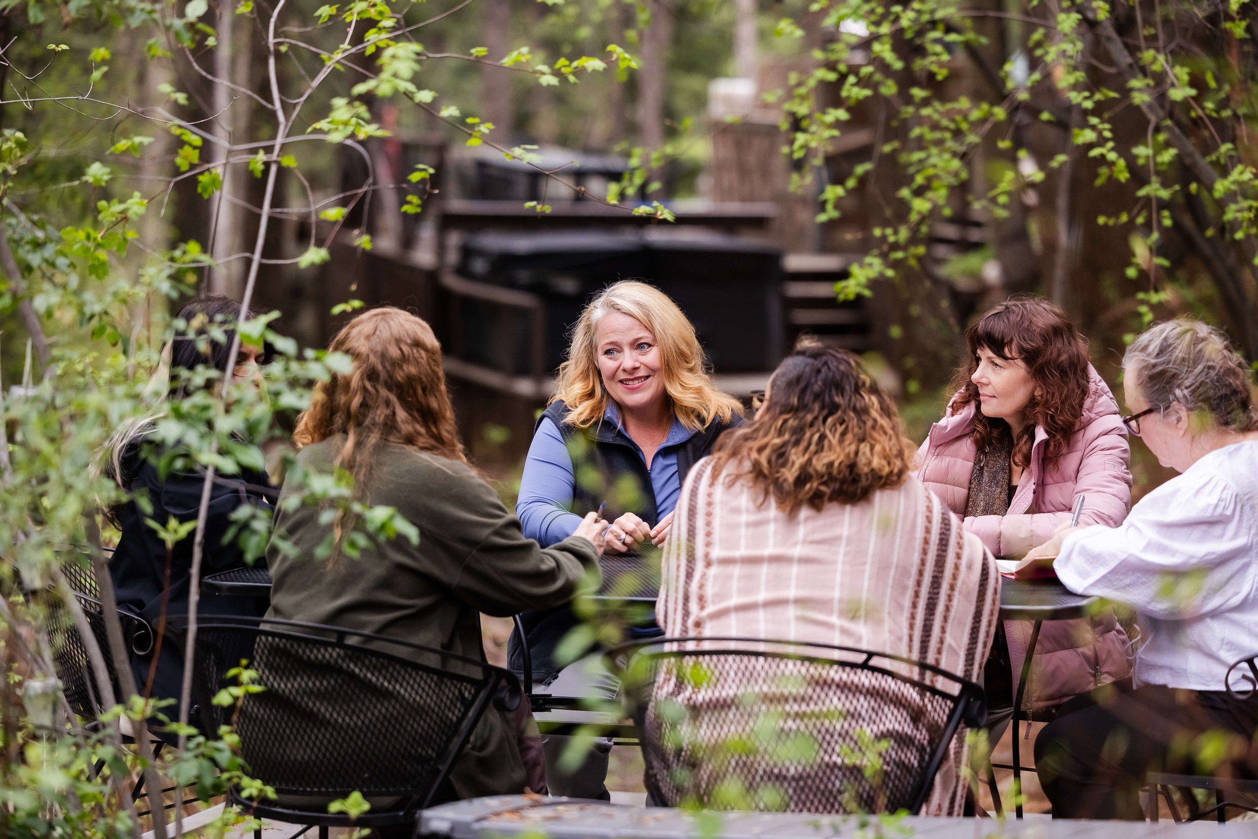 A group of women sitting around a table outdoors in a wooded area, engaged in group coaching at a mountain retreat.