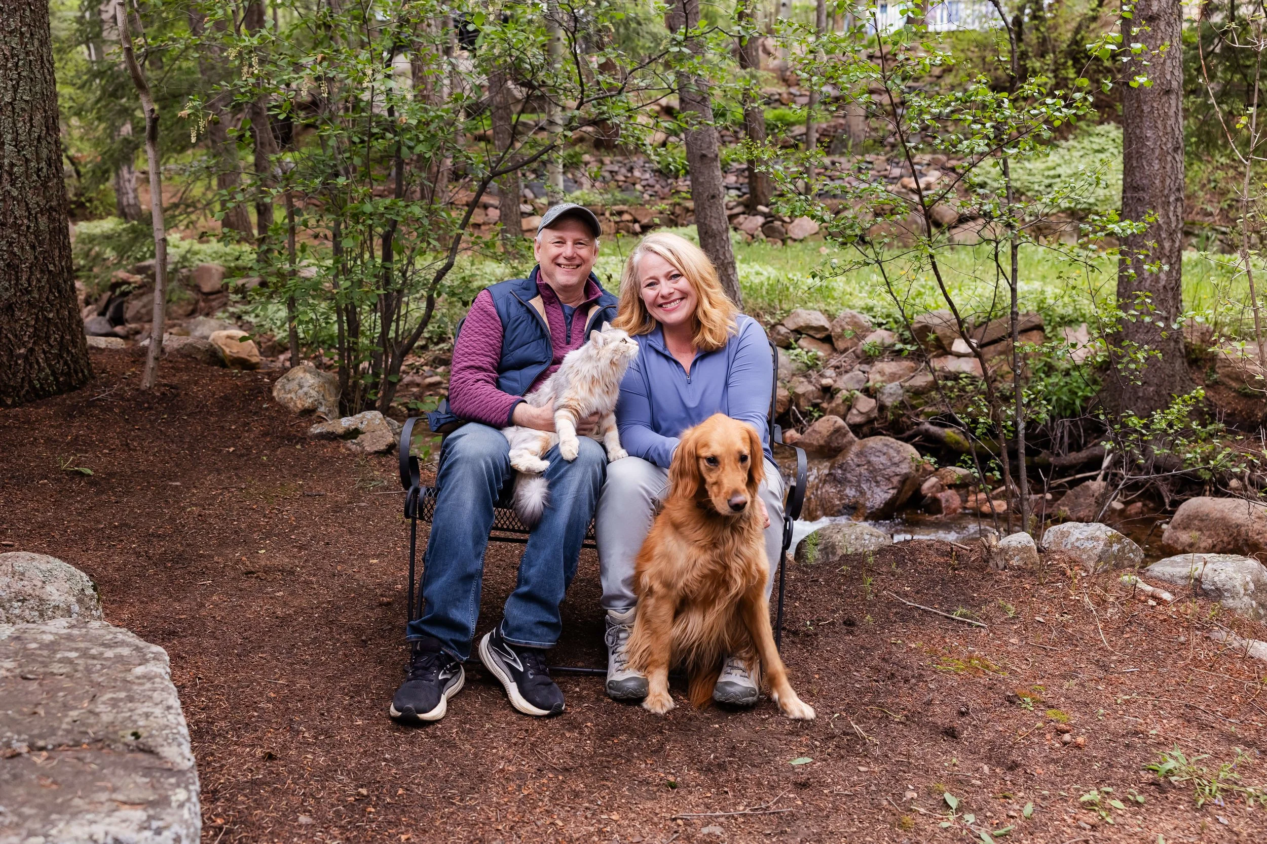 A smiling couple sitting on a bench in Colorado with two dogs, a golden retriever and a cat, during daytime.