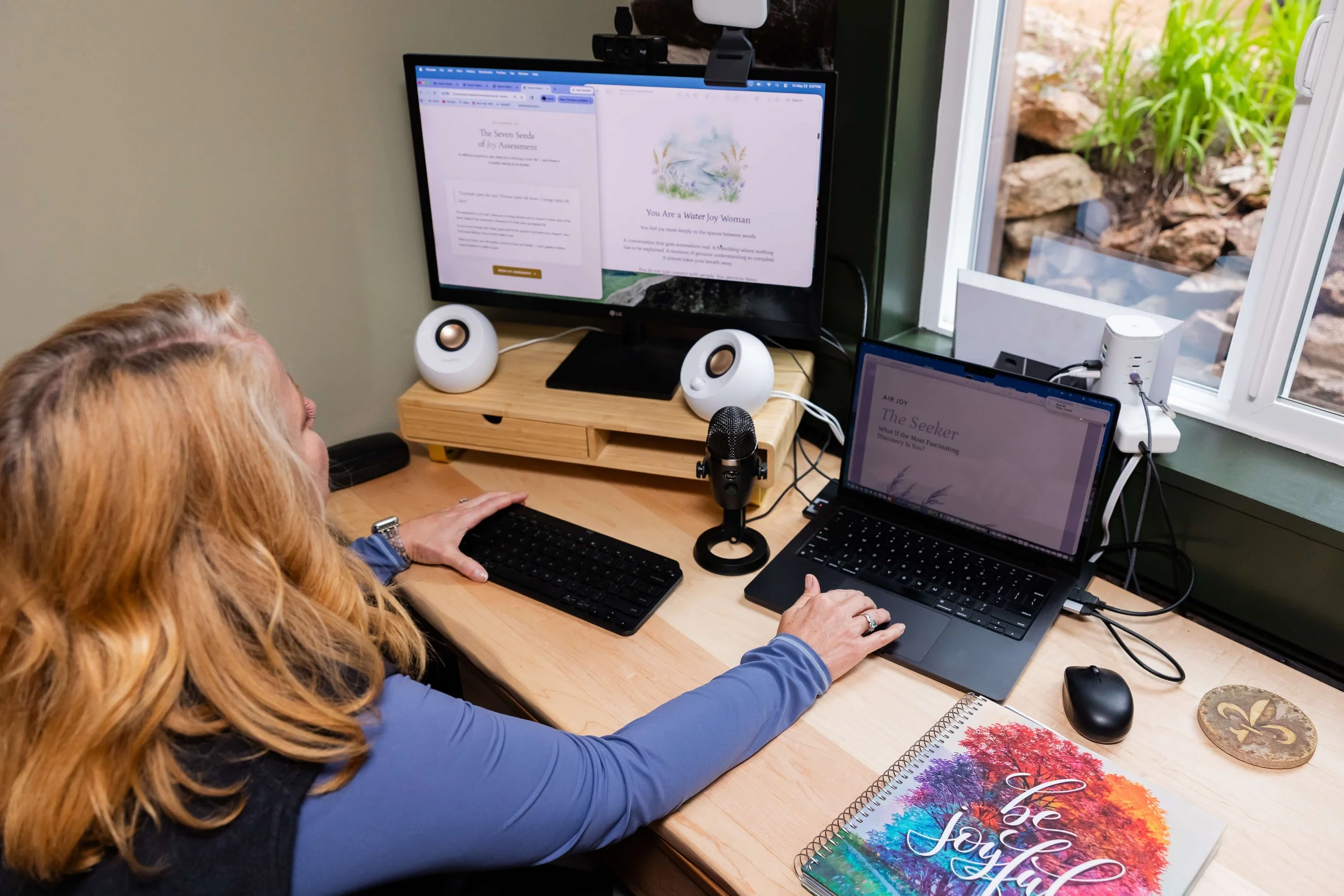A woman is working at a desk with two computers, scheduling individual business coaching in Colorado Springs.