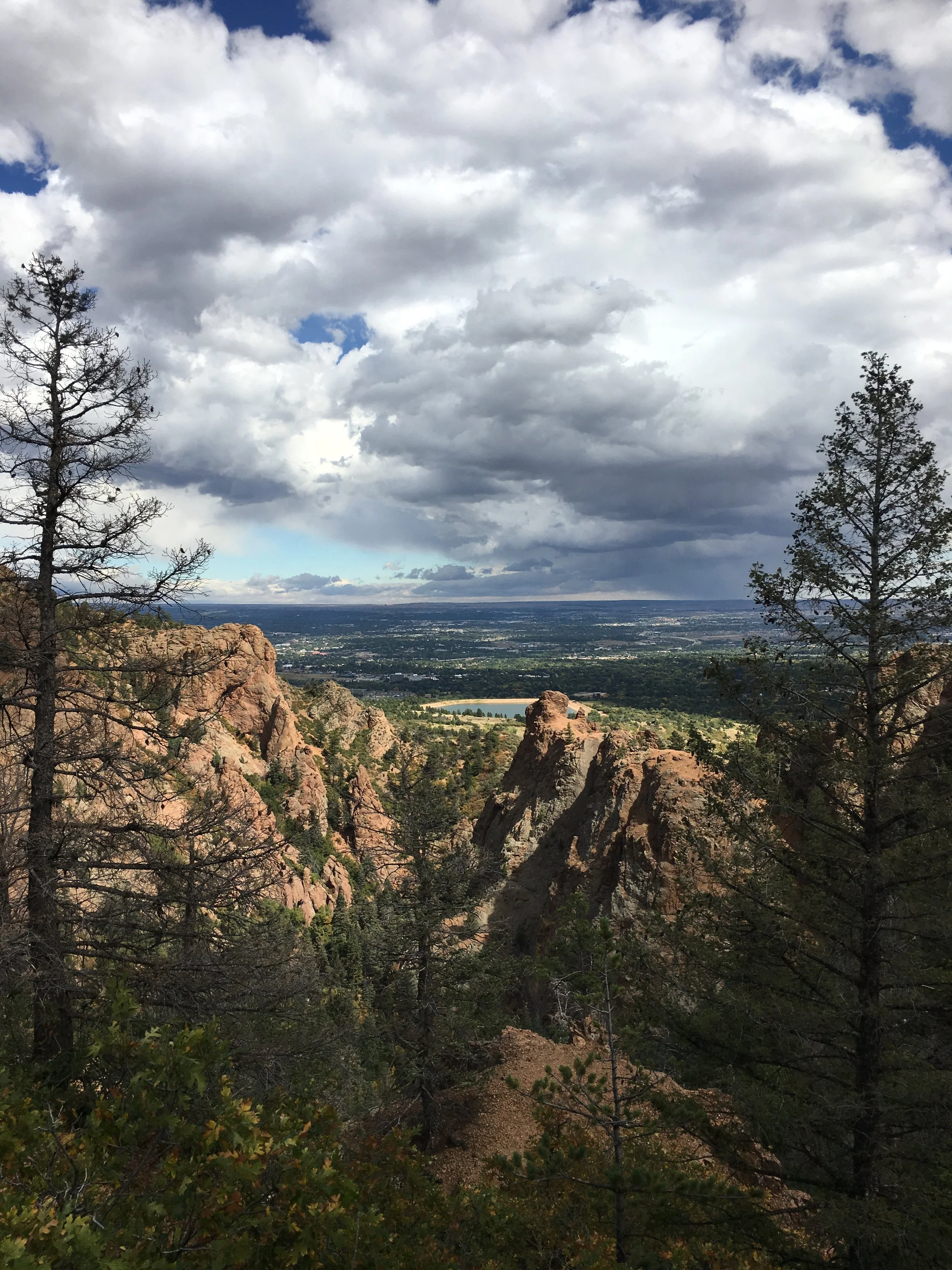 Scenic view of the Colorado mountains with trees in foreground and cloudy sky above, overlooking a city and distant water body.