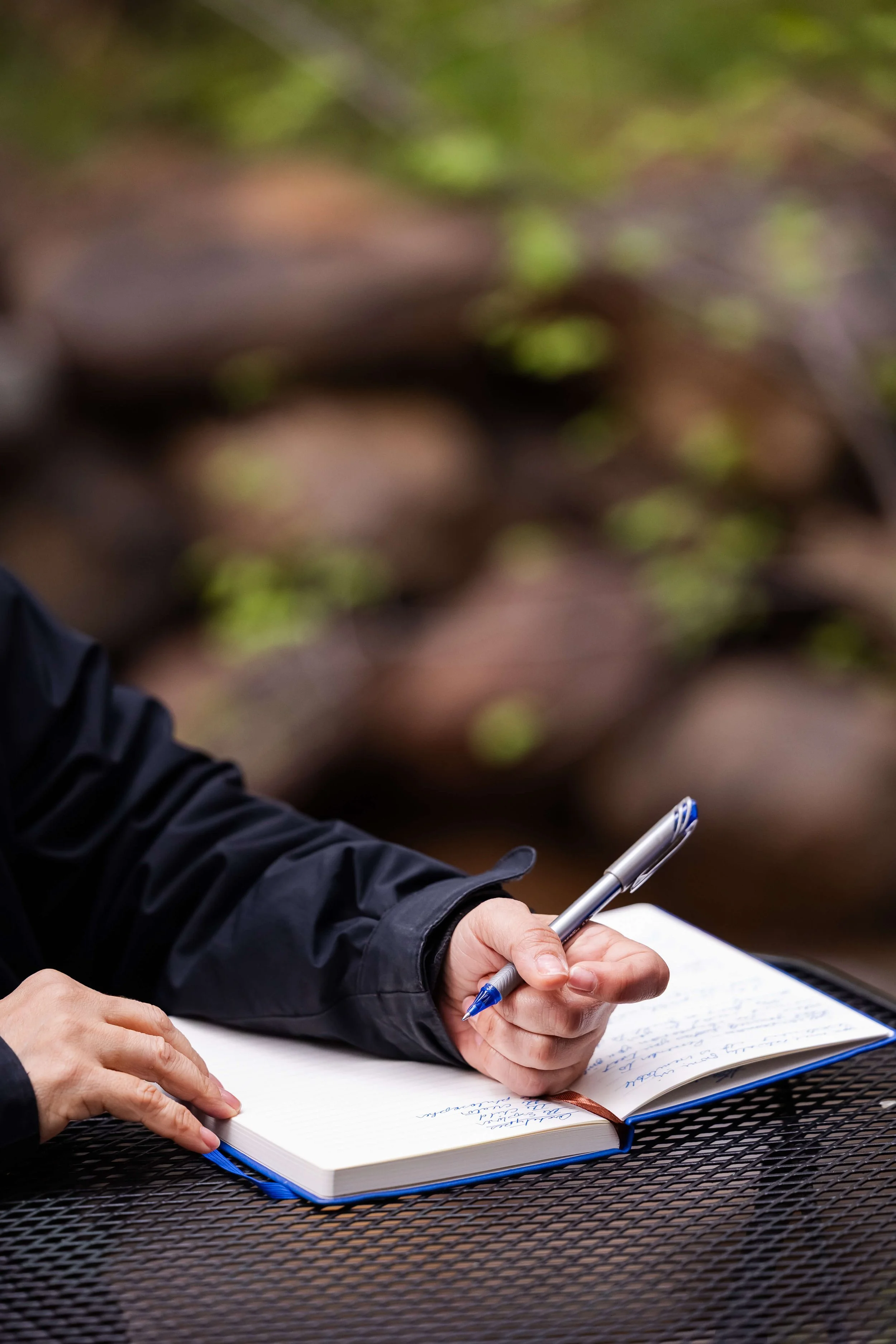 Person's hand holding a blue pen and writing in a notebook on a black metal outdoor table, with blurred green and brown background.
