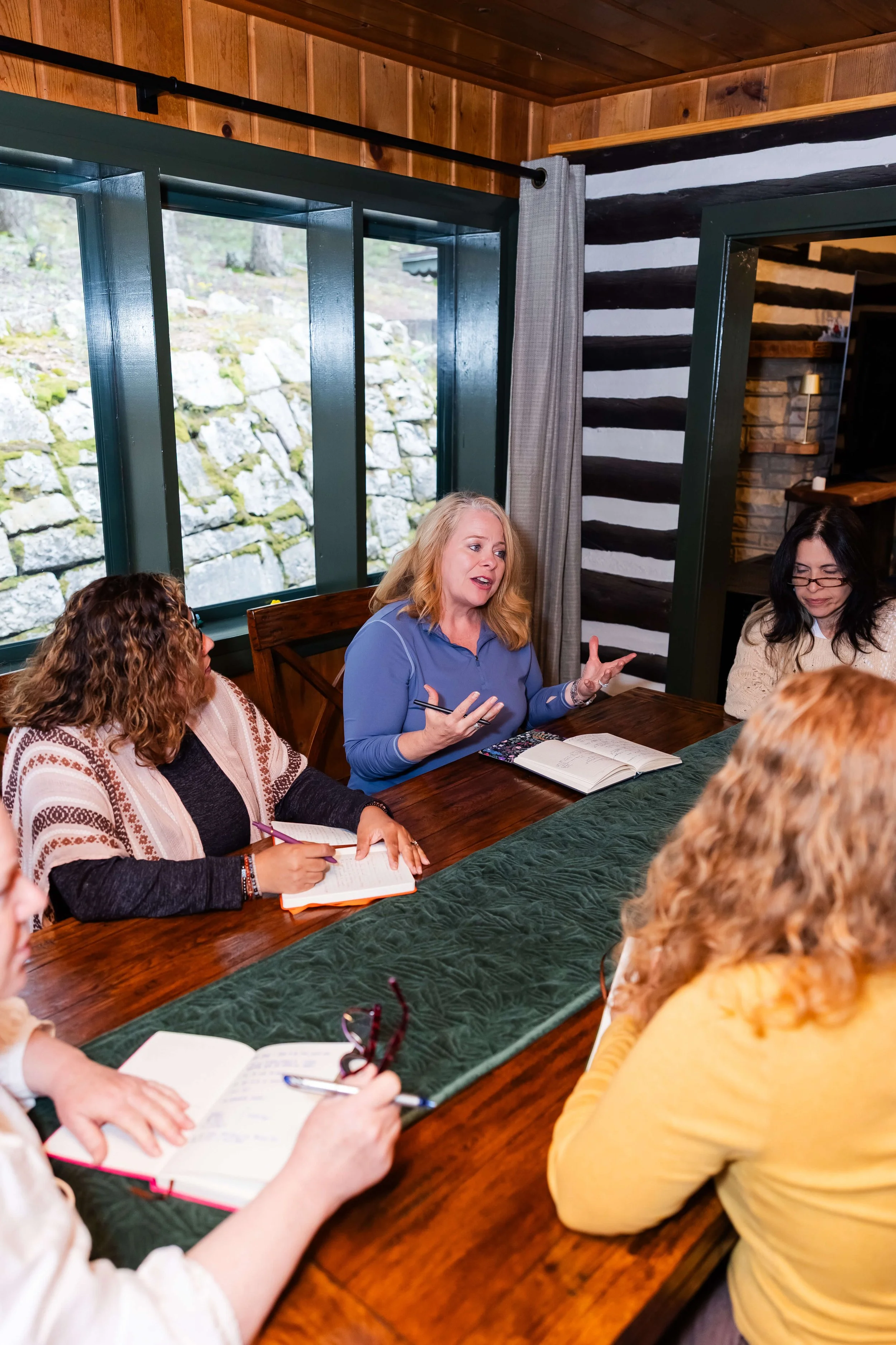A group of women sitting around a wooden table in a cabin with wood-paneled walls and large windows. They are engaged in nonprofit leadership training