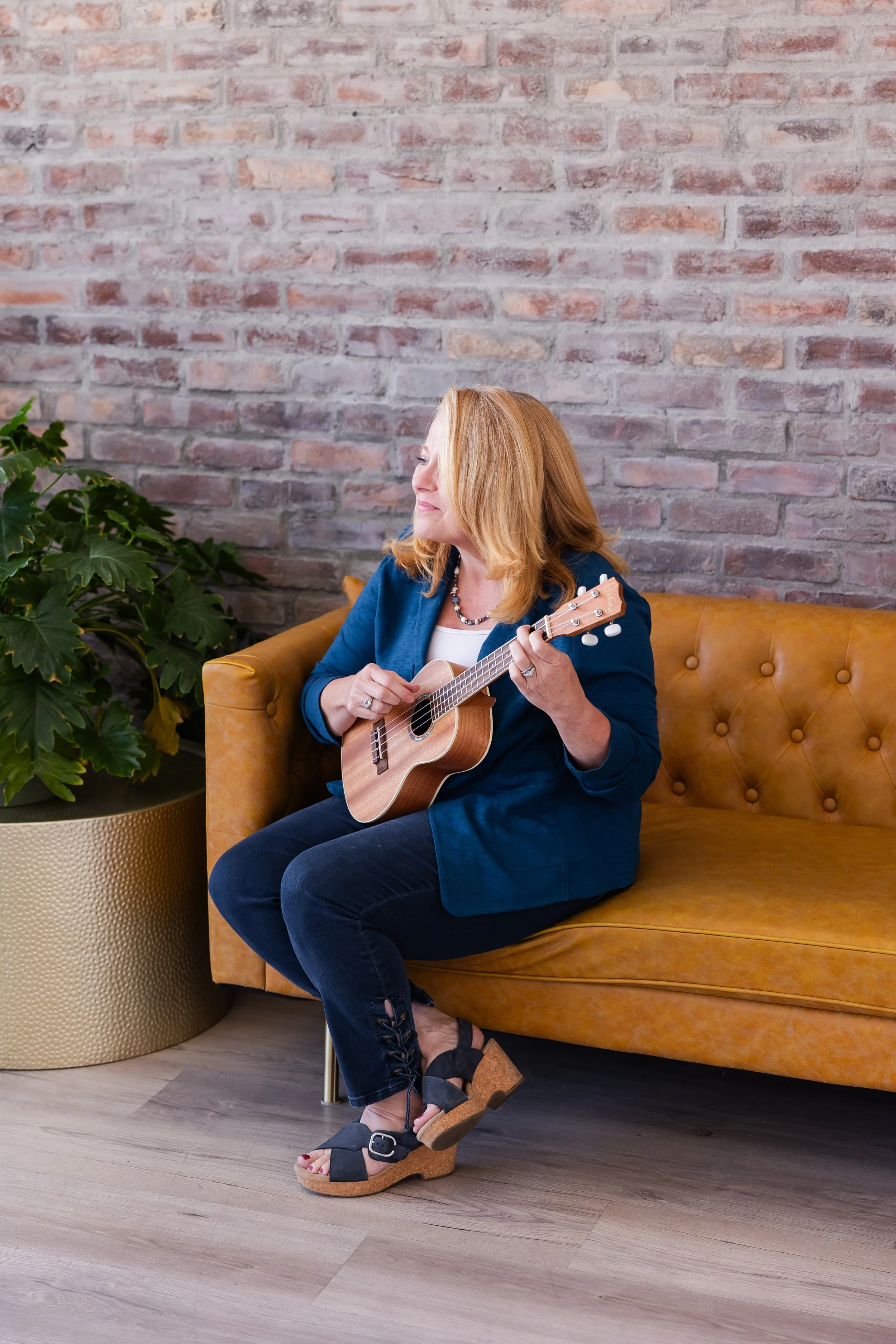 A woman with red hair sitting on a tan leather sofa, playing a ukulele with a brick wall in the background and a large green plant nearby.