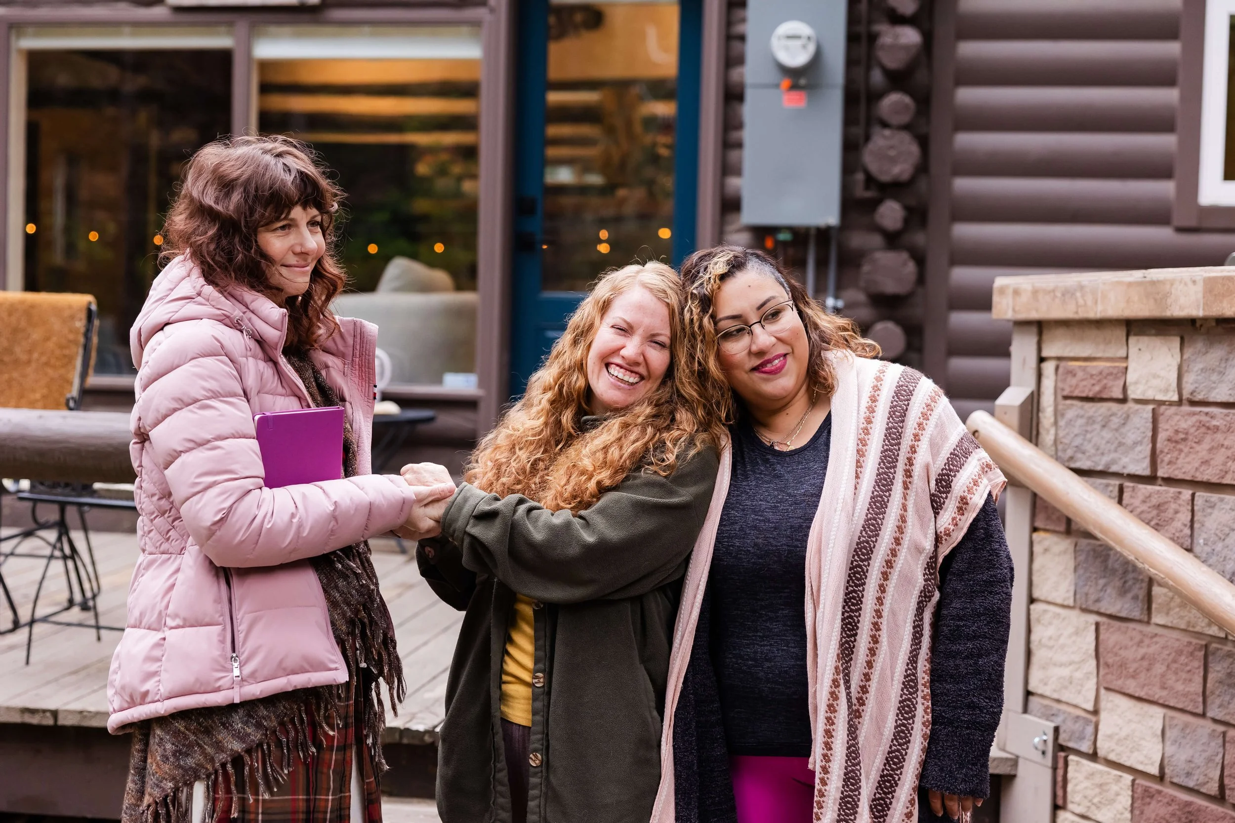 Three women are standing outside on a porch, smiling and holding hands. They are standing in front of a Colorado mountain cabin.