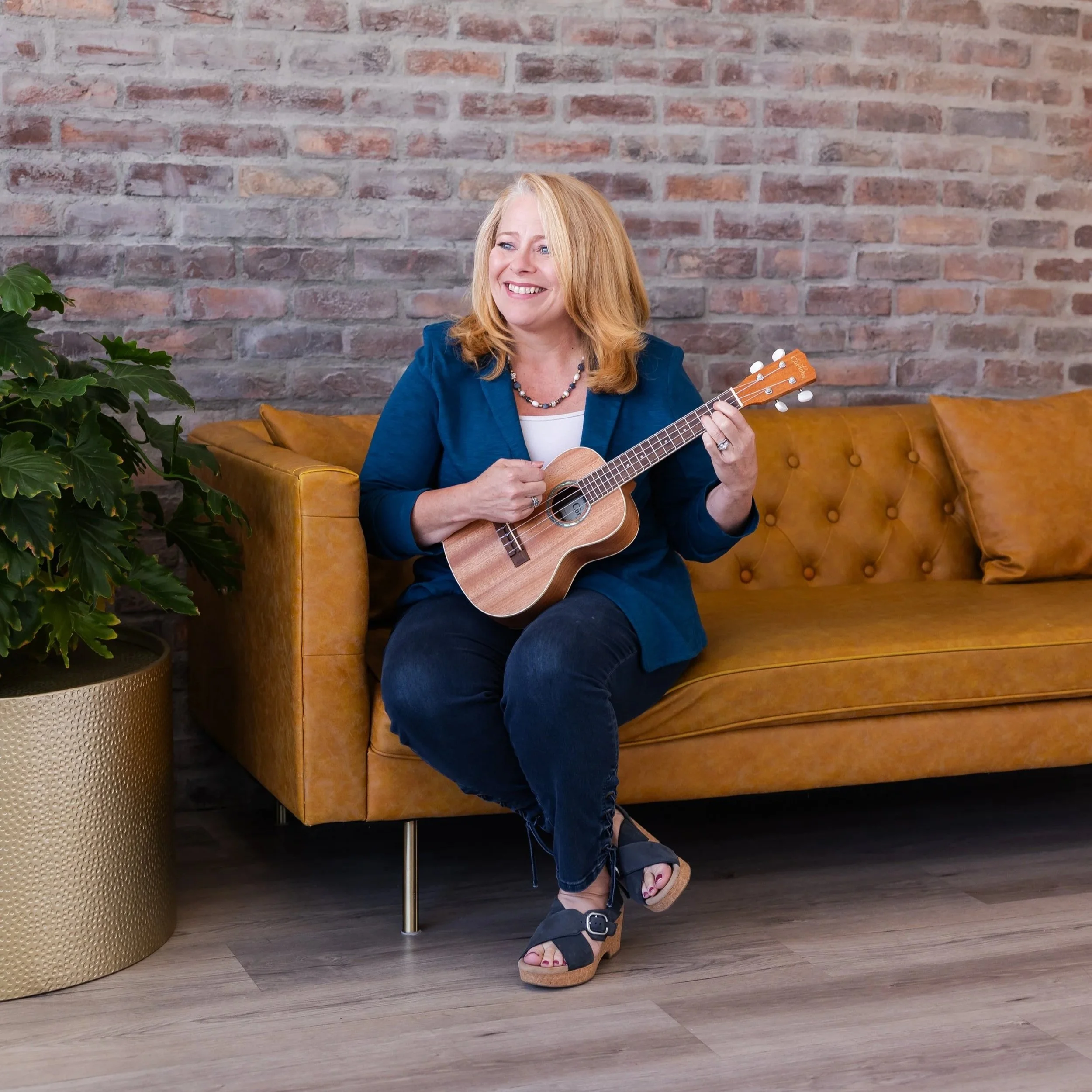 A woman with blonde hair smiling, sitting on a yellow leather couch, holding a small ukulele, wearing a blue blazer, dark socks, and platform sandals, in front of a brick wall with a green plant on her left.
