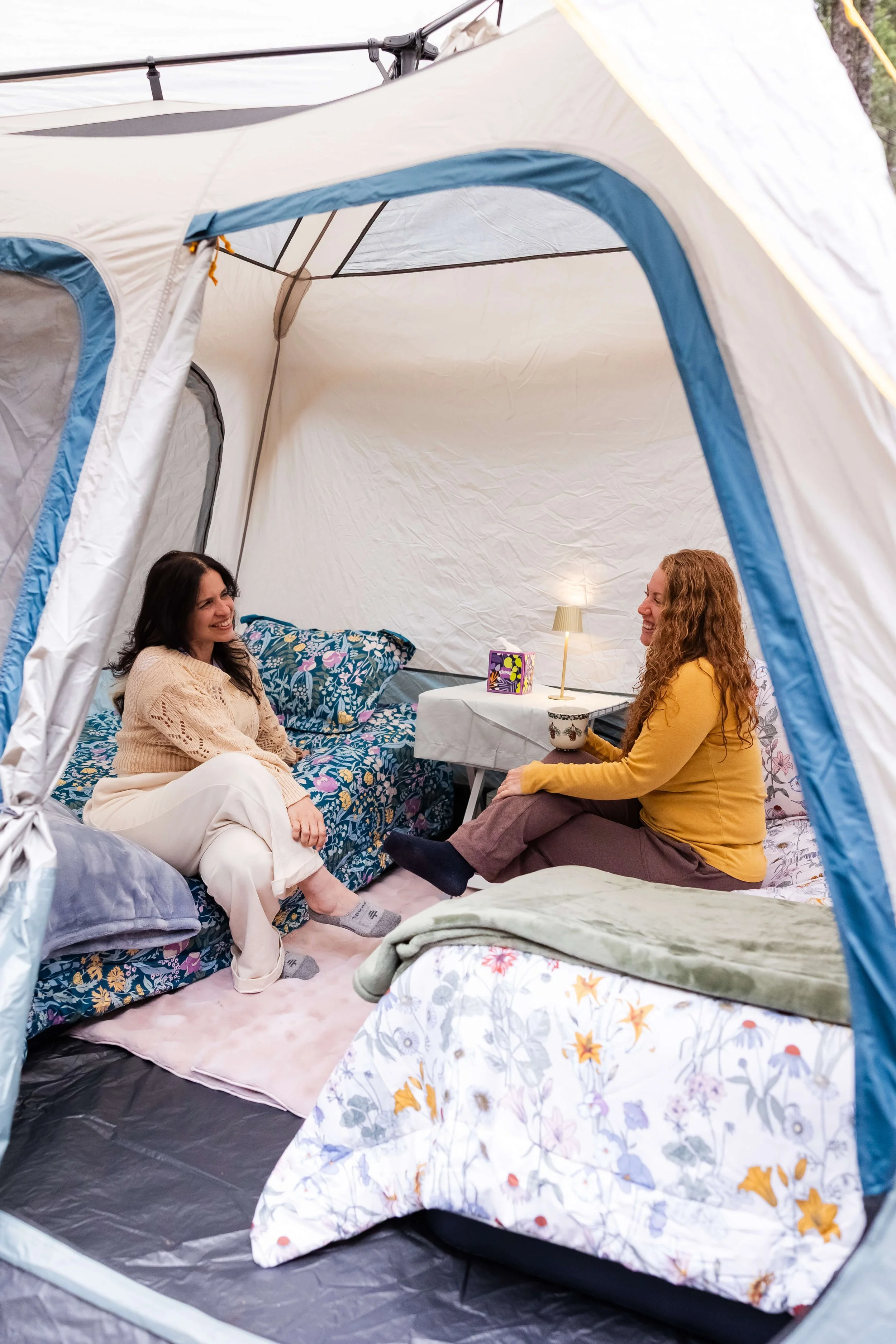 Two women sitting inside a tent, on a nonprofit leadership retreat. The tent has floral bedding, a small lamp, and a box of snacks on a table between them.