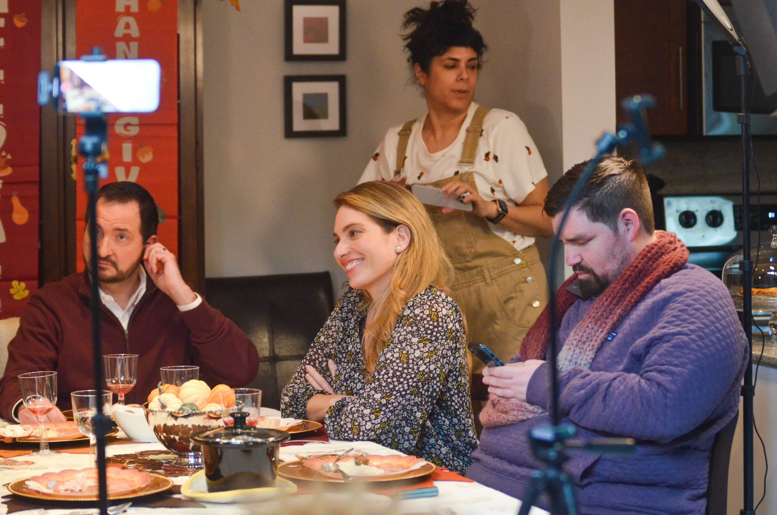 A woman standing behind three people seated at a table, with a laptop, glasses, and a bowl of decorative pumpkins on the table. The woman appears to be overseeing or participating in a gathering or event.