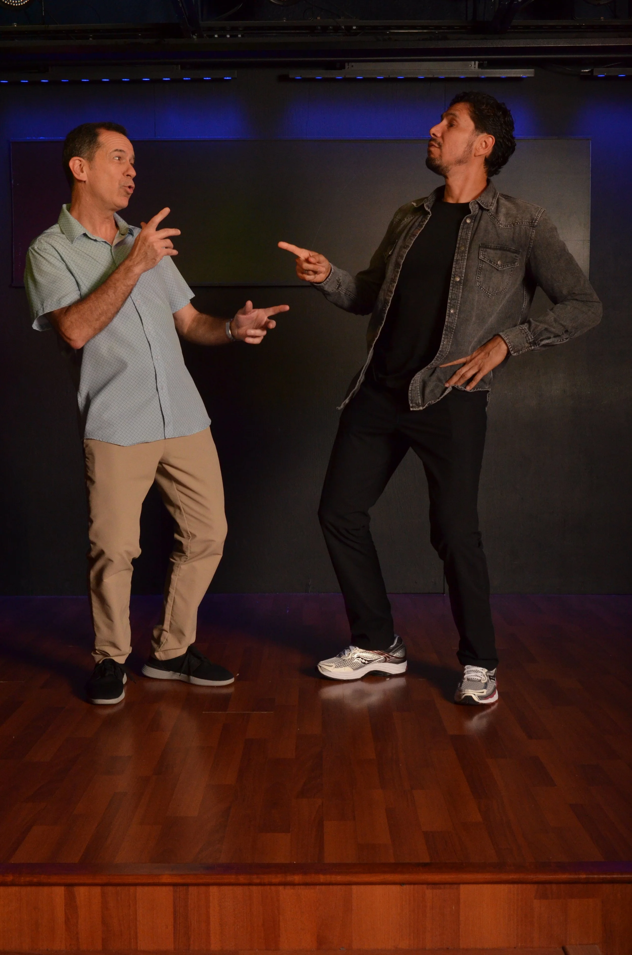 Two men are having a conversation on a stage with a dark background. One man is wearing a light green short-sleeve shirt and beige pants, making a gesture with his hands. The other man is wearing a black shirt, gray denim jacket, black pants, and sneakers, pointing and listening attentively.
