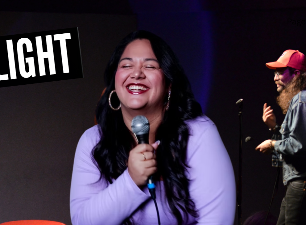 A woman with long dark hair laughing and holding a microphone, wearing a lavender top and hoop earrings, on stage with a man in a cap and denim jacket in the background, and the word 'LIGHT' displayed in large white letters on a black background.
