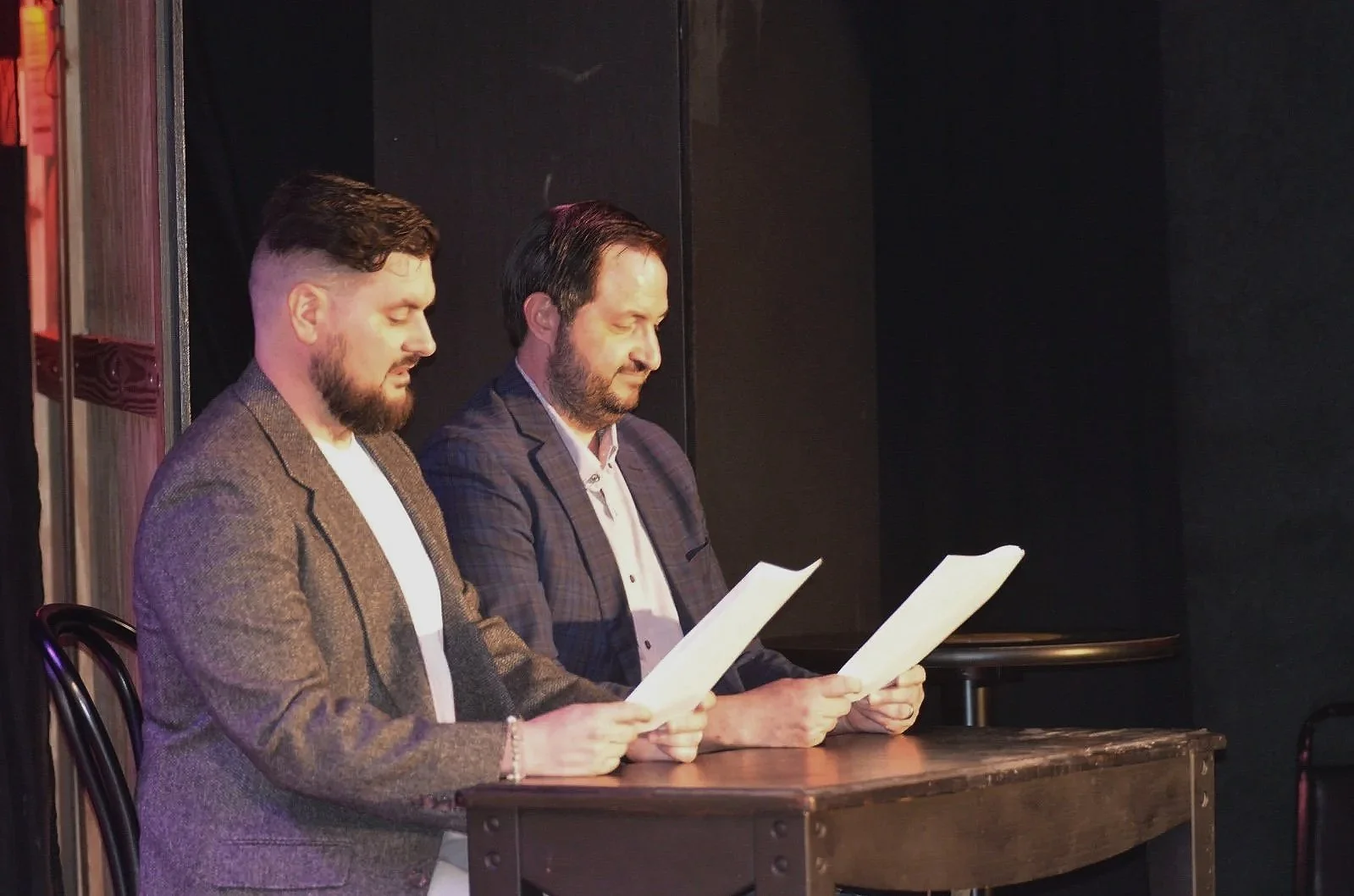 Two men sitting at a wooden table reading papers, dressed in suits, in a dark room or stage setting.