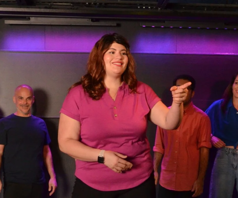 A woman with long, wavy brown hair wearing a pink polo shirt pointing, with three other people standing behind her in a colorful indoor setting.
