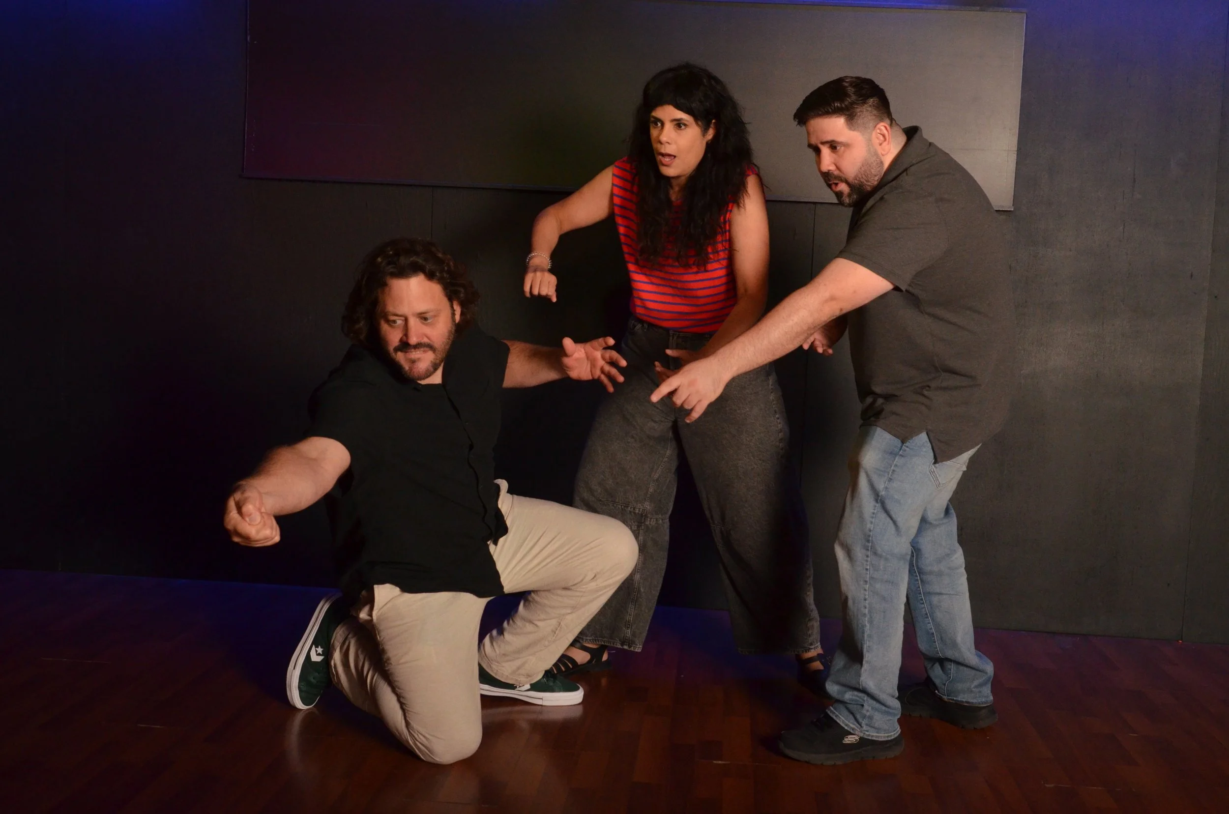 Four people are gathered in a dark room with a black wall. One person is seated on the floor, and three are standing around, with one of them pointing at the seated person. They appear to be engaged in conversation or an interaction.