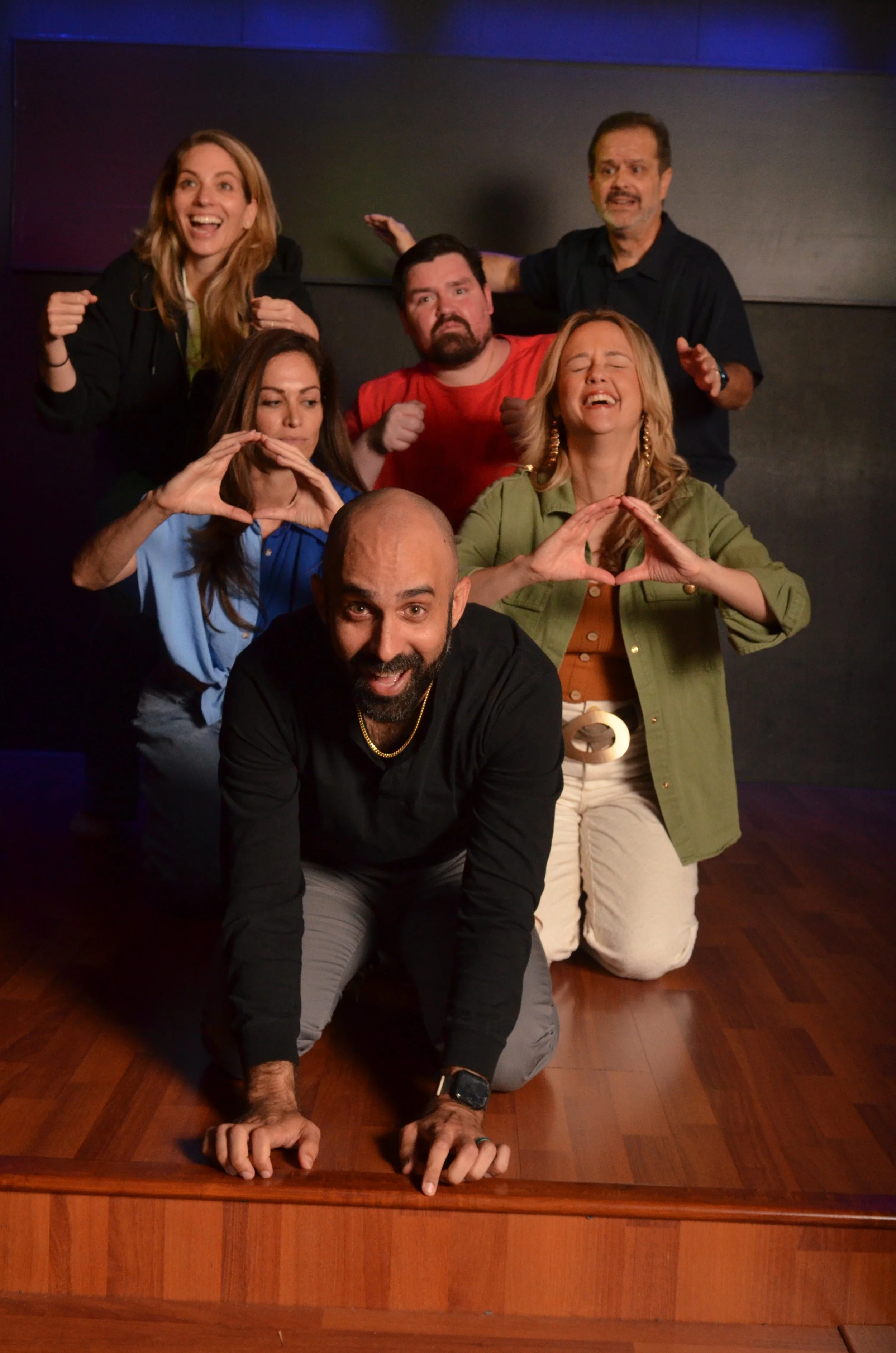 Six people posing for a mirror selfie, two women, three men, and one person whose face is not visible, with smiling, serious, and playful expressions, standing and kneeling on a wooden floor in an indoor setting.