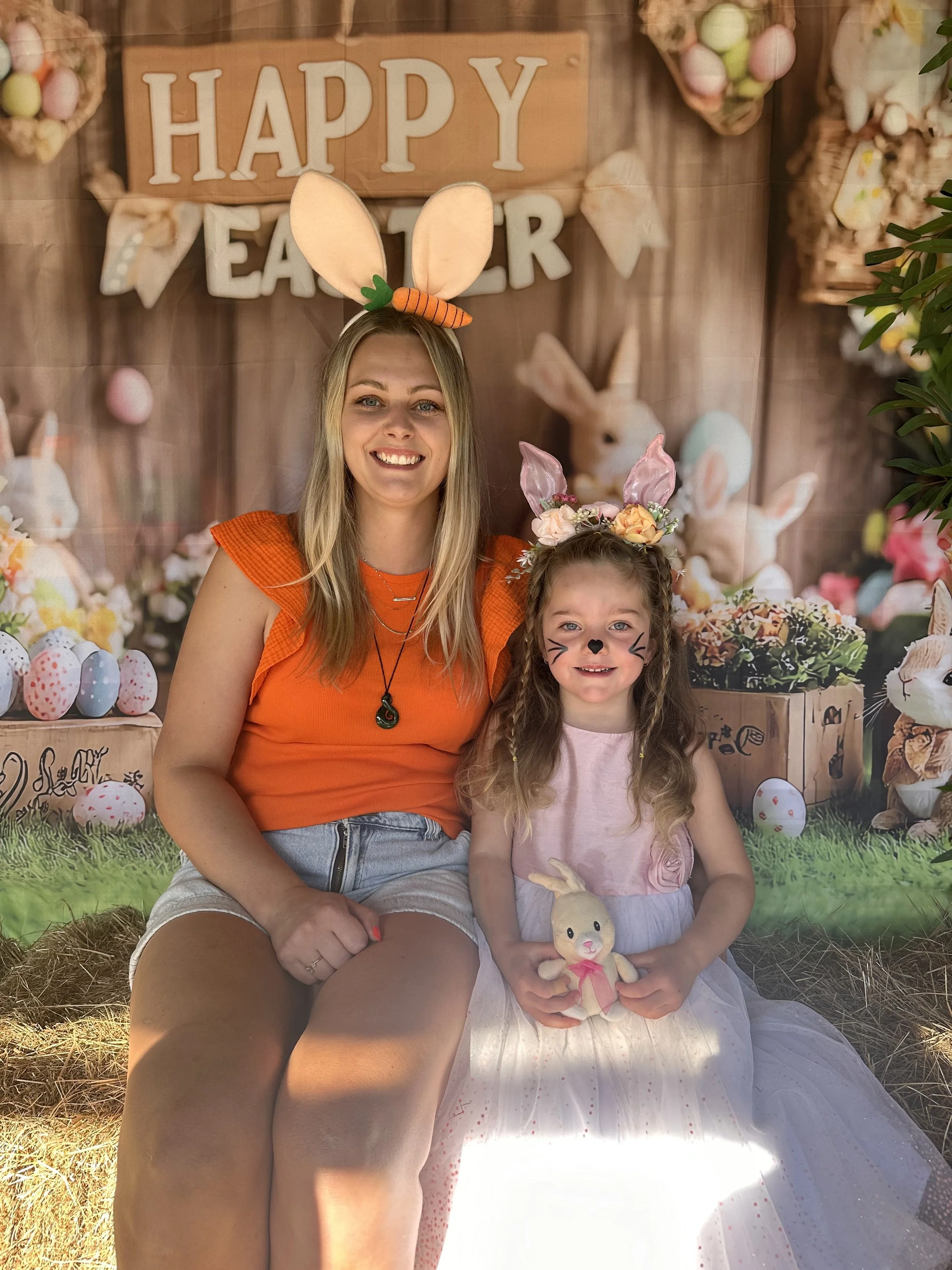 A woman and a girl dressed in Easter-themed costumes, sitting on hay with a decorated backdrop that has a 'Happy Easter' sign and Easter decorations.