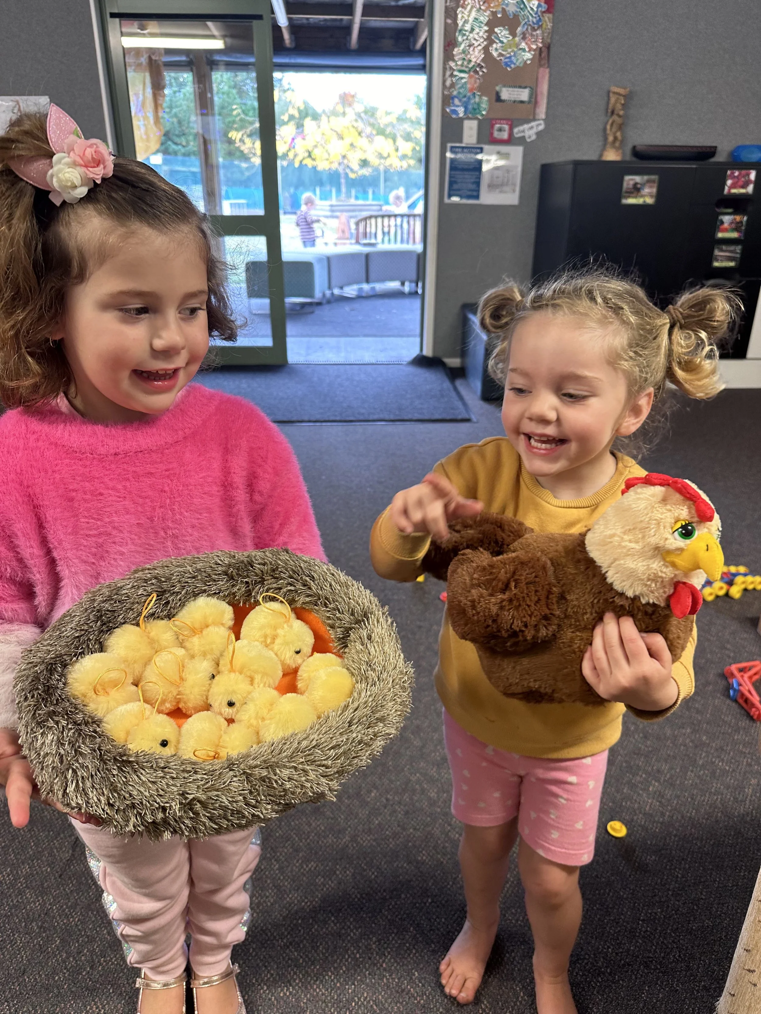 Two young girls standing indoors, each holding a plush toy chicken—one yellow and fluffy, the other brown with an embroidered face and red comb. The girl on the left is wearing a pink fuzzy sweater, pink pants, and sandals, with a pink bow headband a