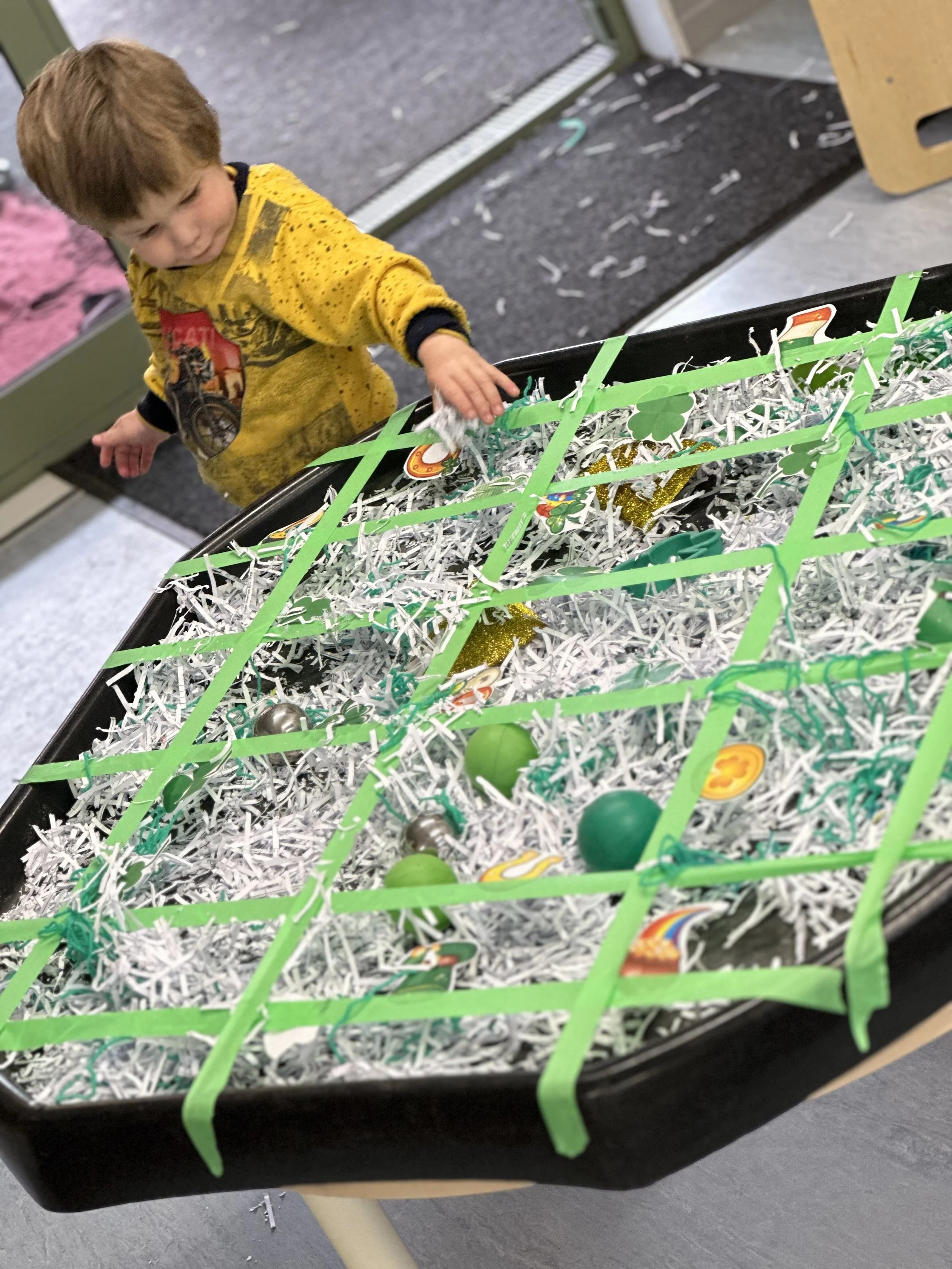 A young boy wearing a yellow long-sleeve shirt is reaching into a black container filled with shredded paper and plastic eggs, decorated for Easter.