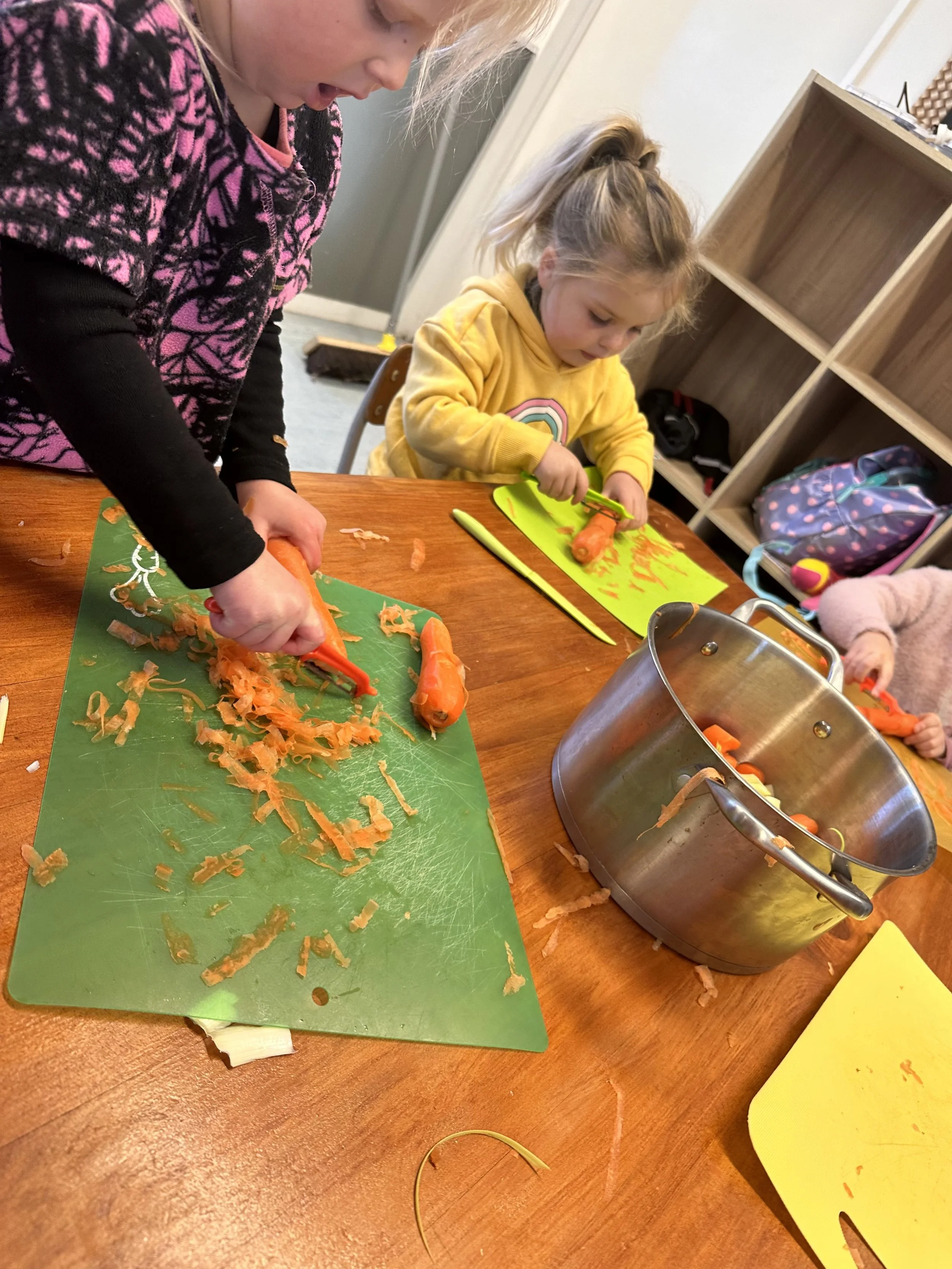 Child and an adult slicing carrots on cutting boards at a wooden table in a classroom or kitchen setting.