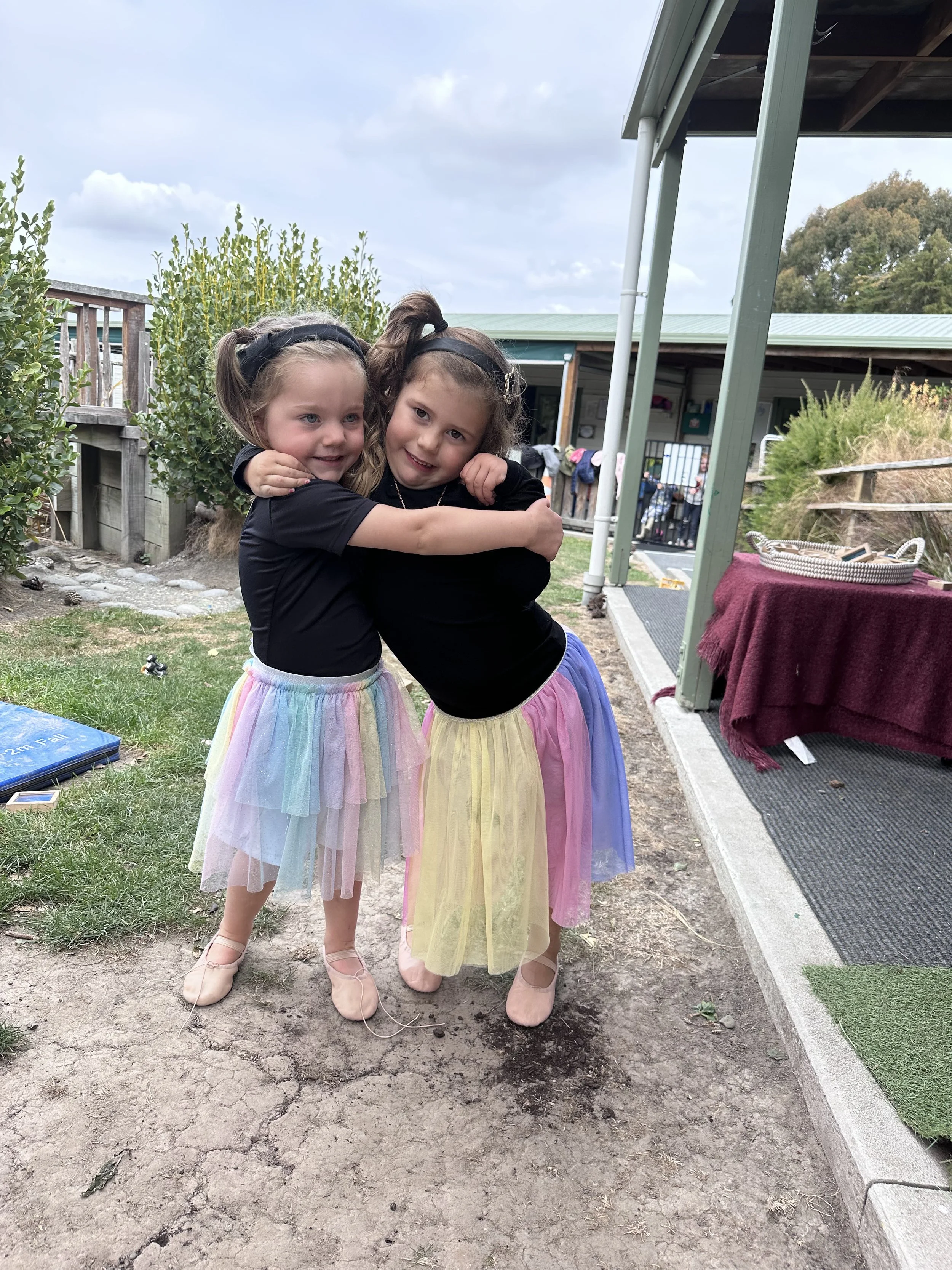 Two young girls with long hair, wearing matching black tops and colorful skirts, hugging each other and smiling outdoors.