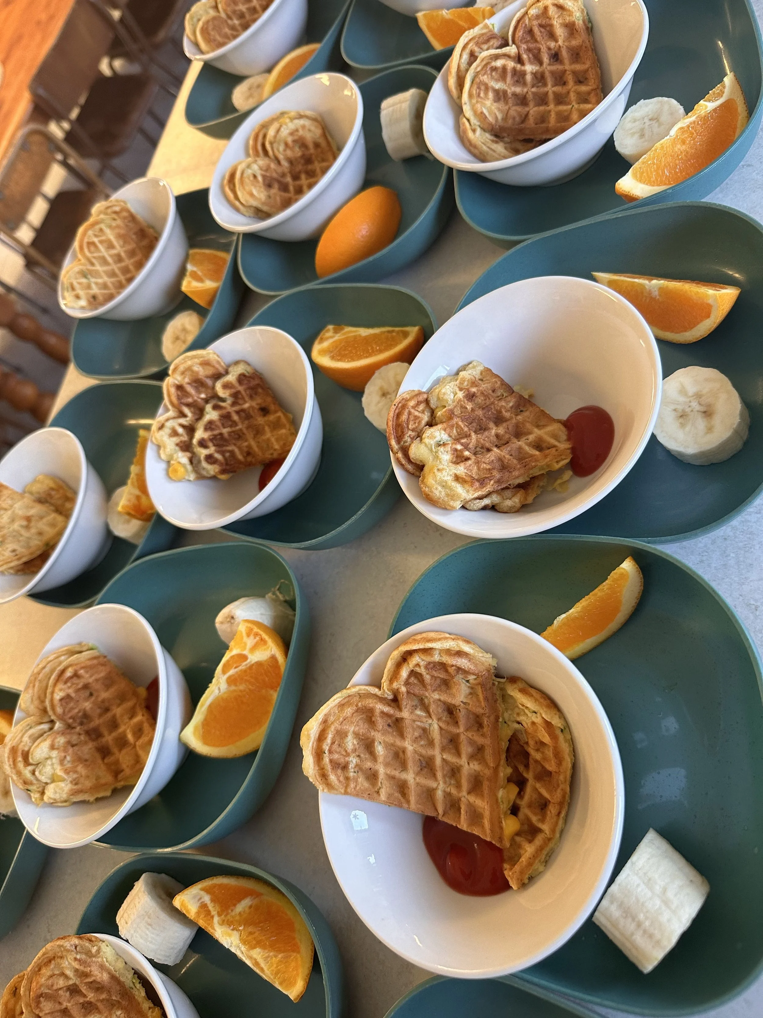 Multiple blue trays with white bowls containing waffles, orange slices, banana slices, and small marshmallows on a table.